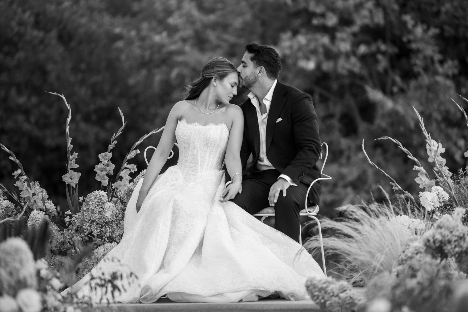 A romantic black and white wedding photo of a couple embracing among wildflowers and tall grasses in a garden setting.