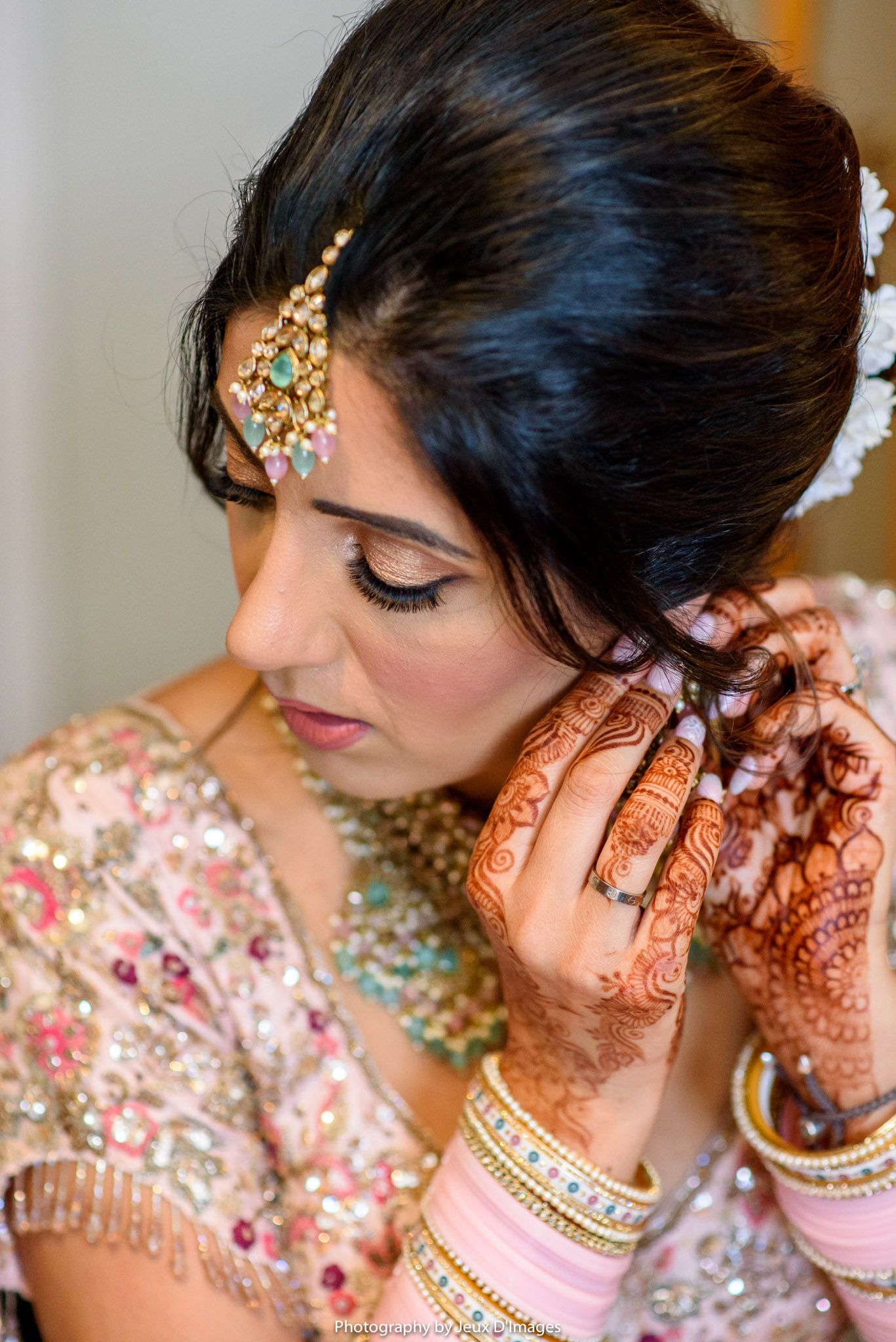 Indian bride adorned with mehndi, bangles, and maang tikka jewelry adjusts her earring in bridal attire.