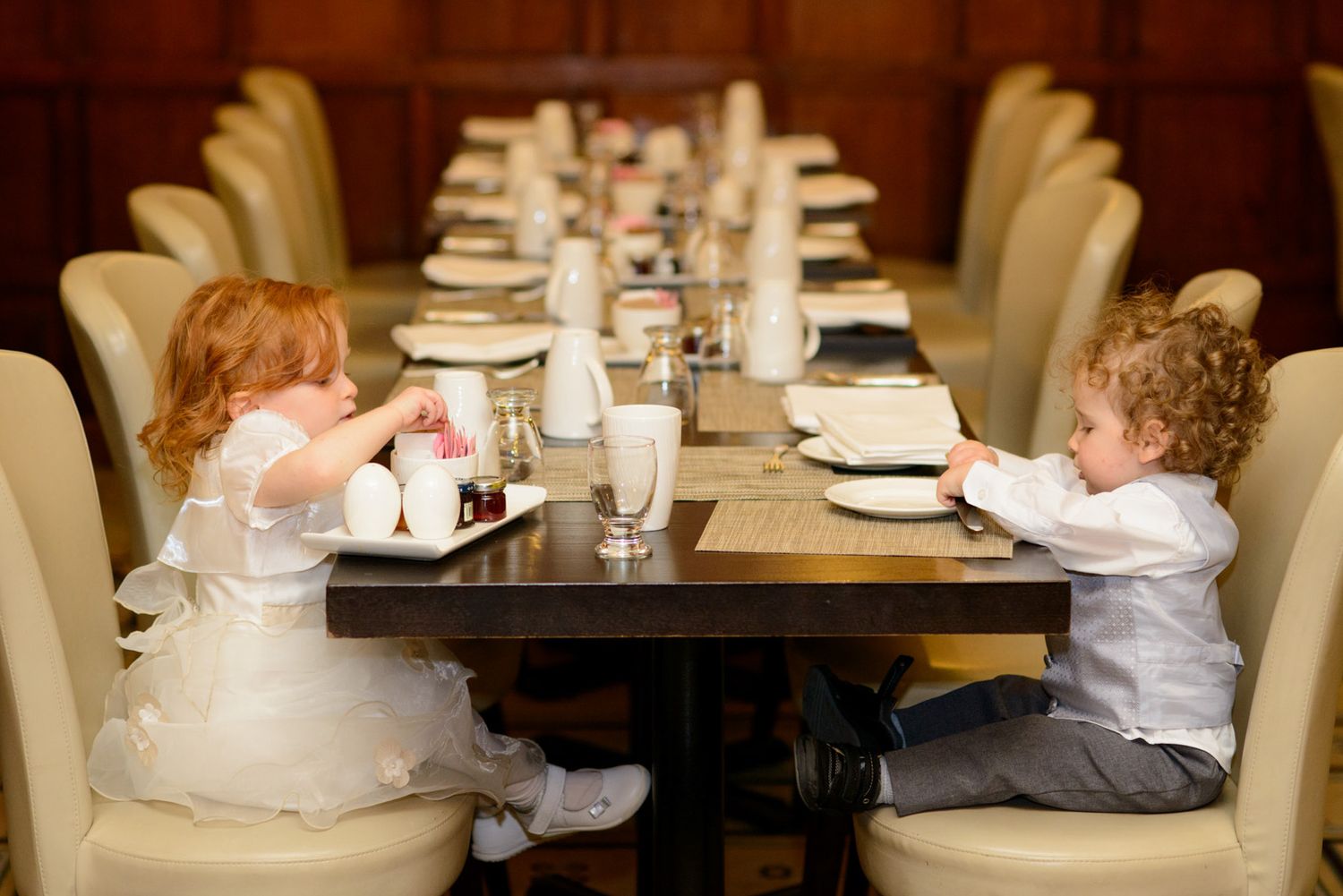 Two young children in formal attire sit across a long elegantly set dining table at a wedding reception.