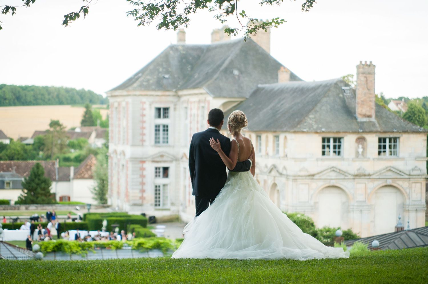 Bride and groom stand arm in arm on a lush lawn overlooking an elegant French chateau wedding venue.