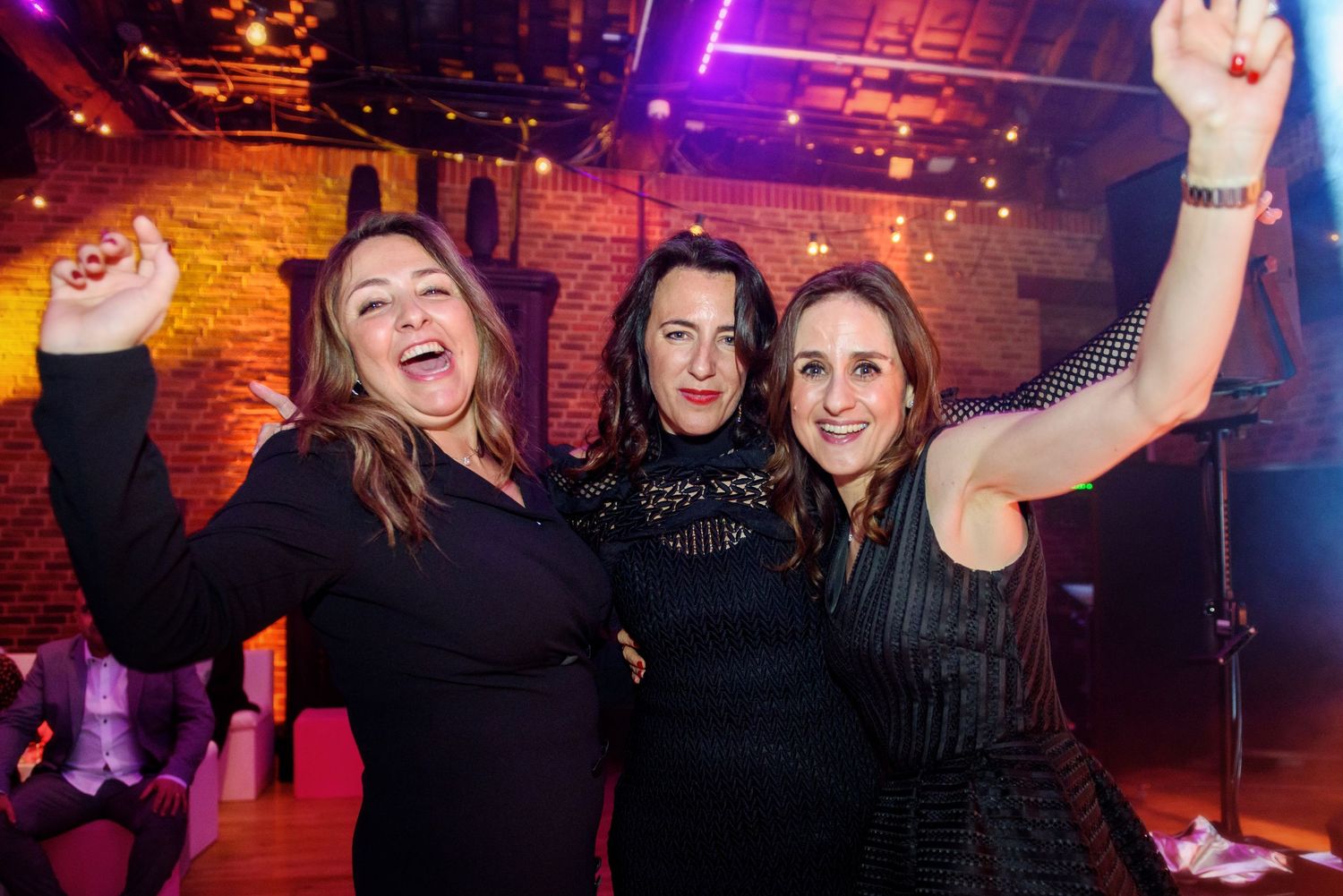 Three friends celebrating and dancing with raised arms at a nightclub with purple lighting and brick walls.