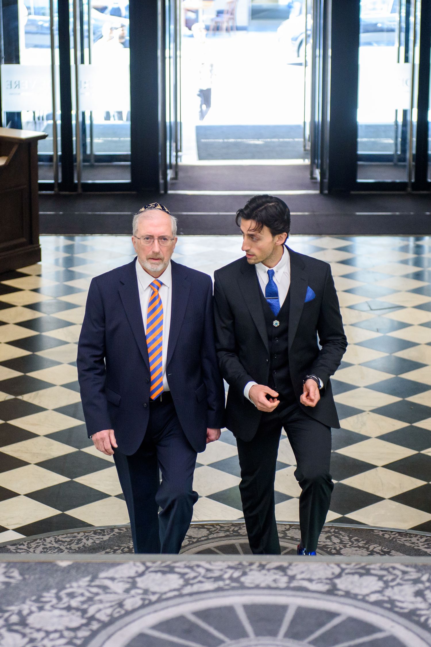 Two businessmen in dark suits having a conversation while standing on a checkered floor in an elegant hallway.