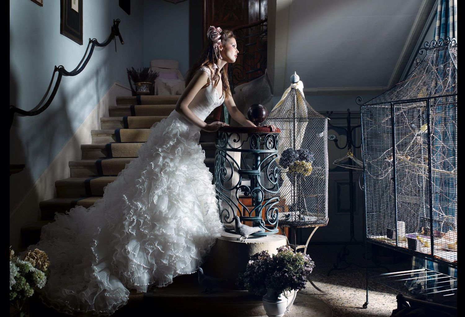Bride in white gown poses gracefully on ornate staircase beside vintage birdcage in elegant historic interior.