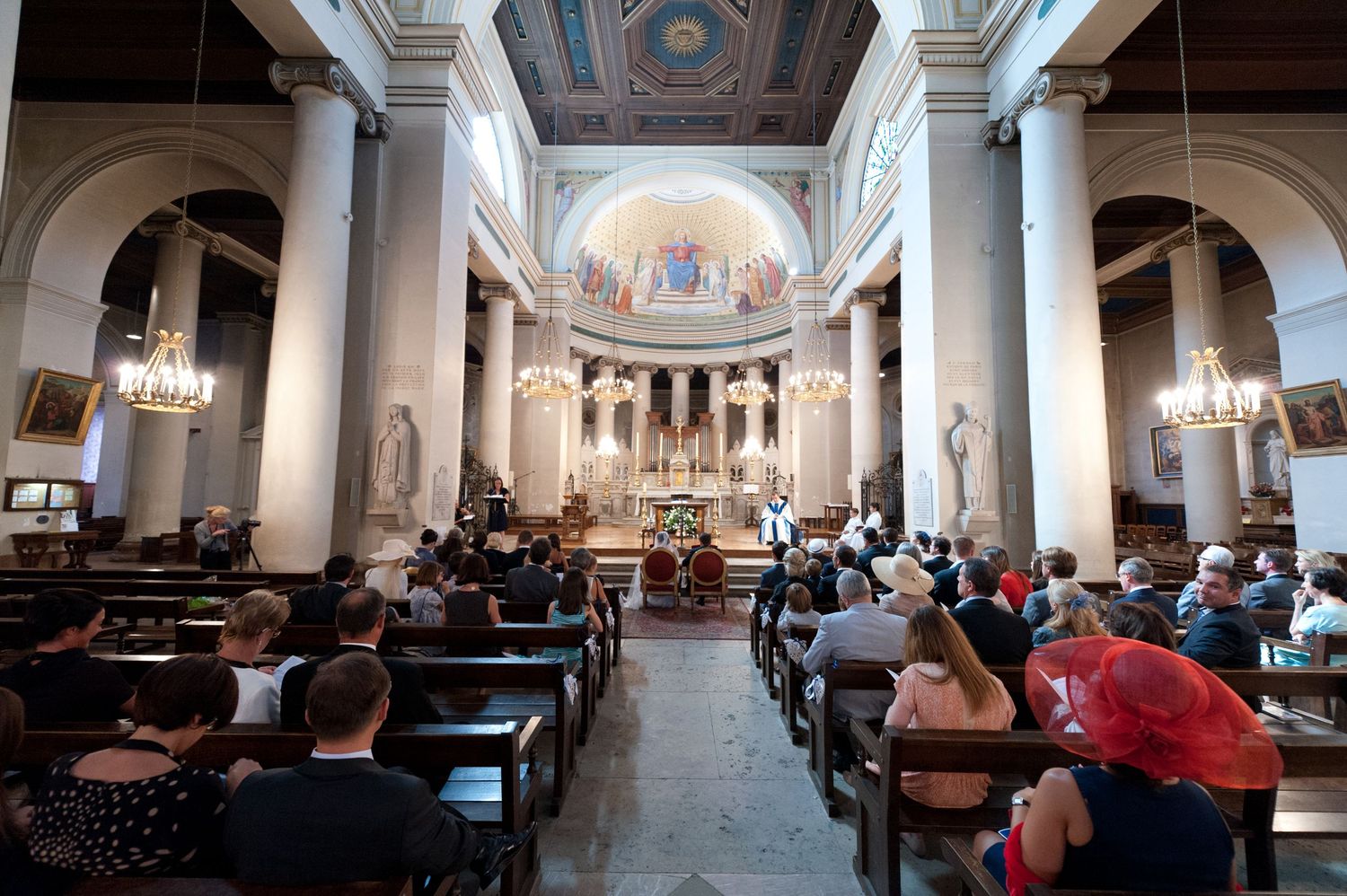 Congregation gathered inside an ornate neoclassical church with tall columns, chandeliers, and a decorated altar during a service.