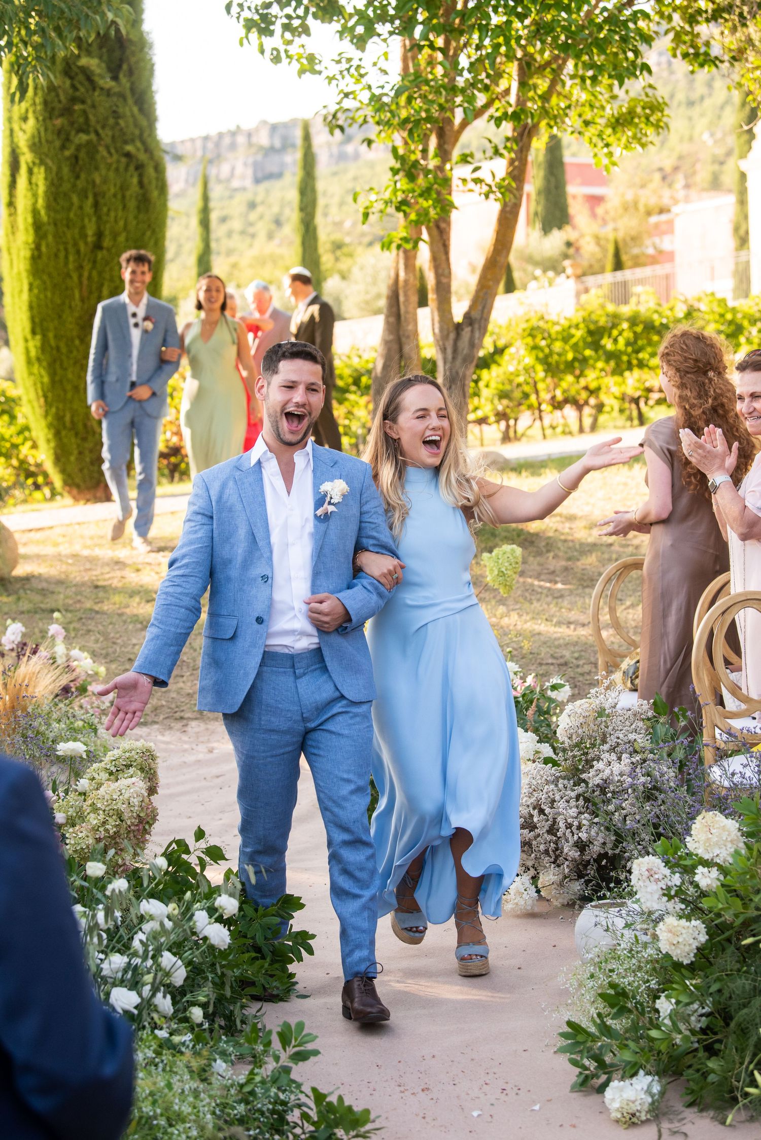Wedding party members in light blue suits joyfully celebrate while walking down a garden path lined with white flowers.