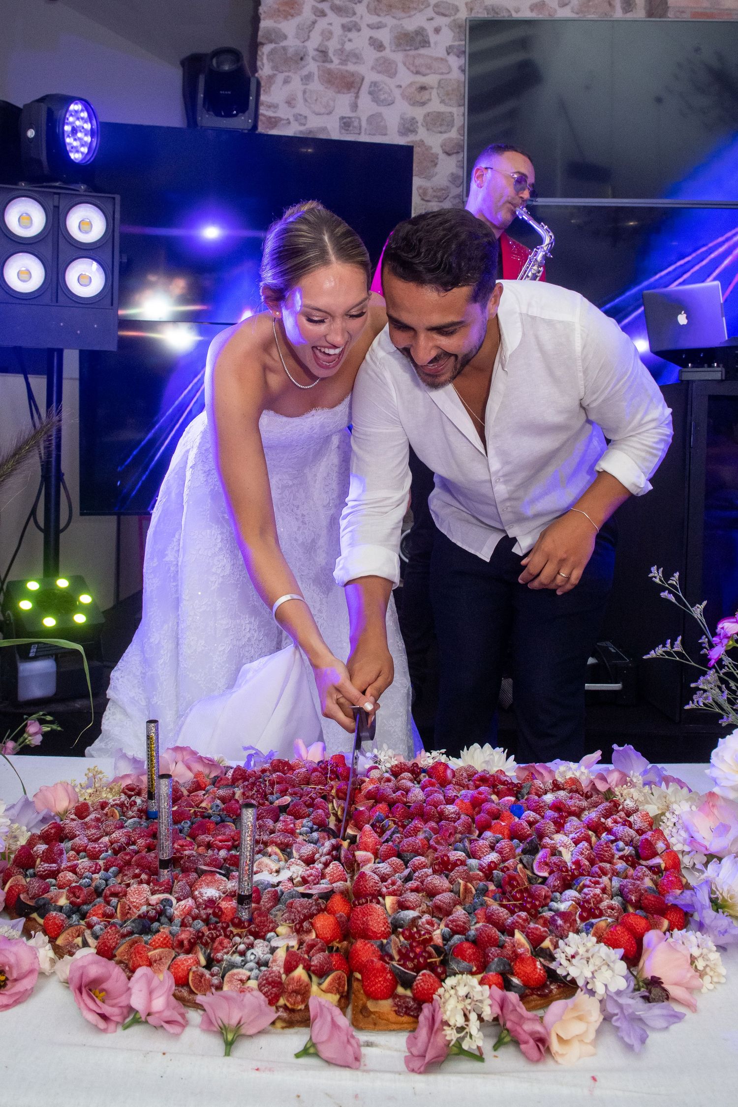 A couple in formal attire cuts into a stunning display of red and pink berries and flowers at a celebration.