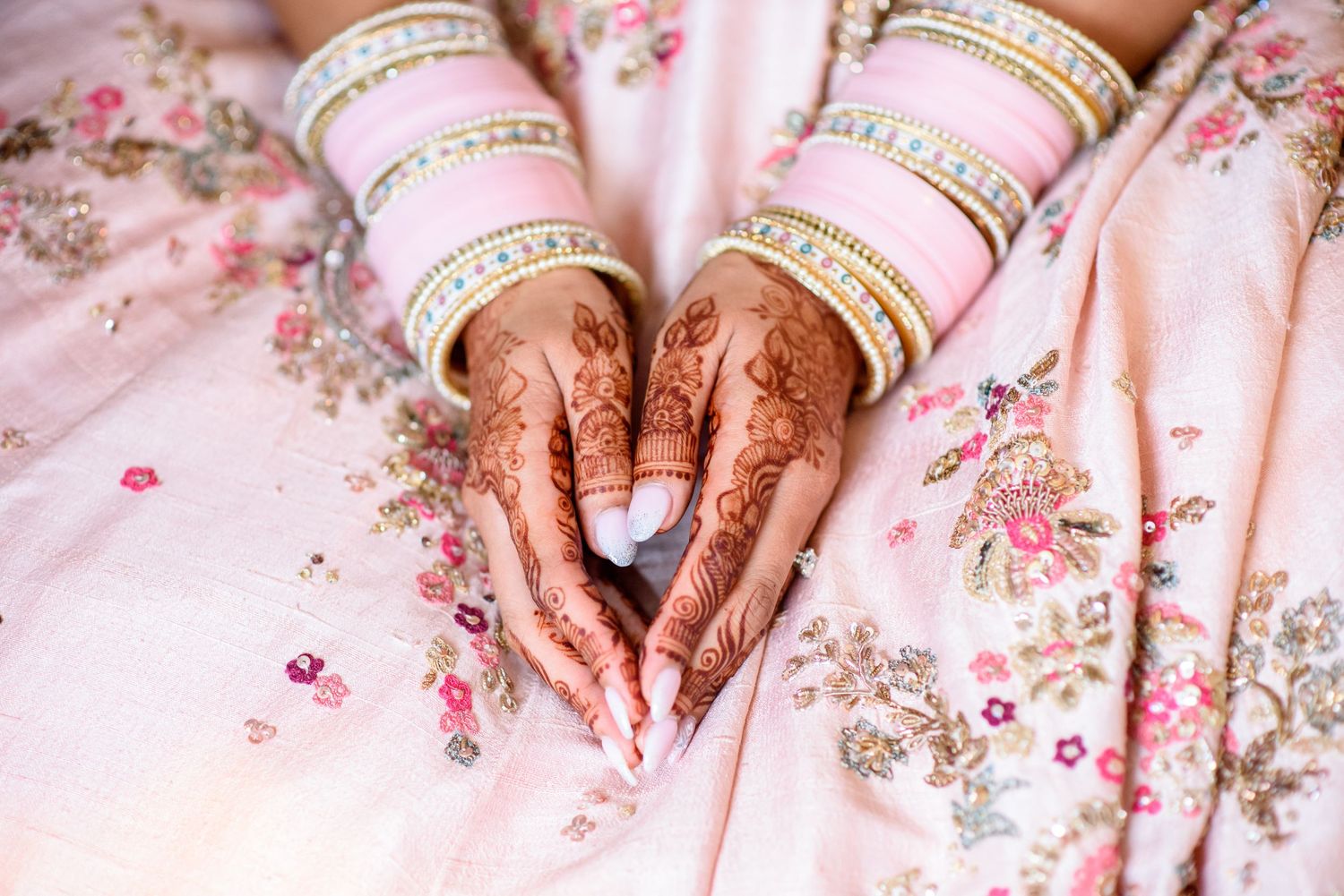 Pink and gold bangles adorned with intricate henna designs rest on floral fabric in a traditional Indian wedding setting.