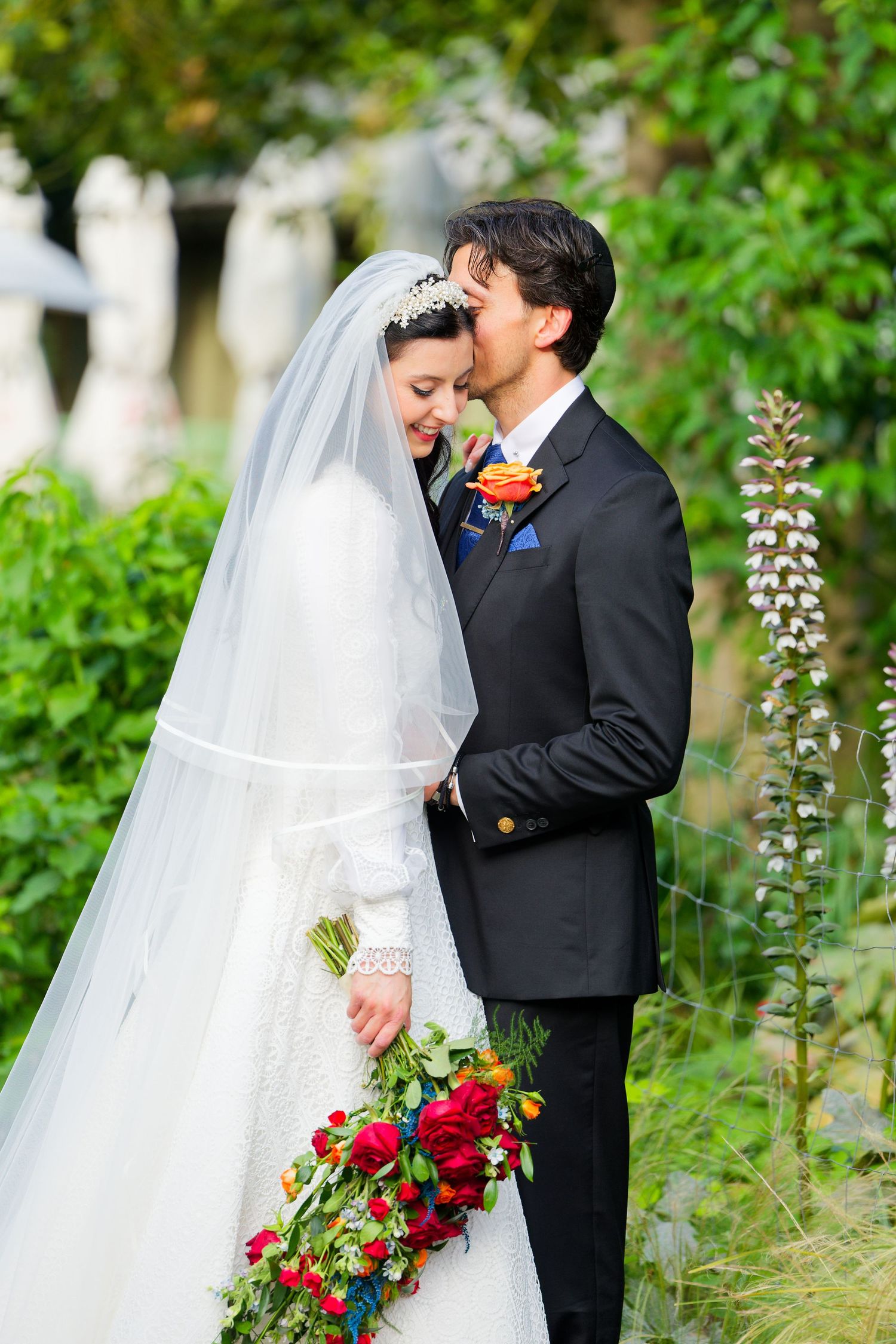A romantic wedding couple shares tender moments in a garden setting with vibrant red bouquet and flowing white veil.