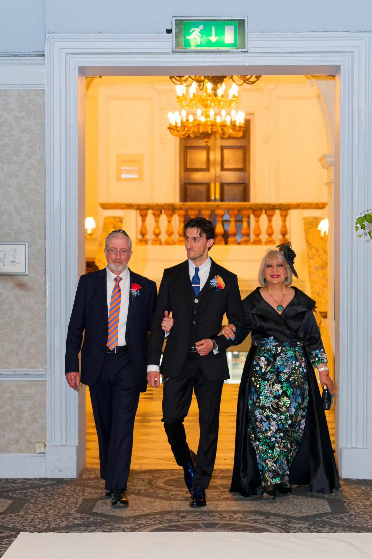 A group of formally dressed people walking through an elegant hallway with chandeliers and ornate decor.