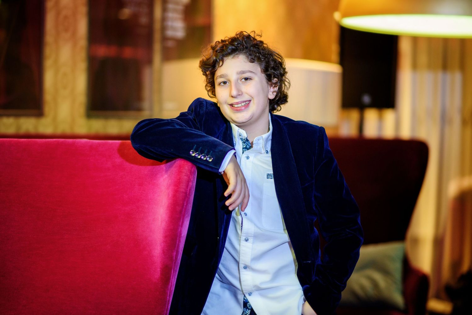 A young person in a navy blazer and white shirt smiles while sitting in a restaurant with red booth seating.