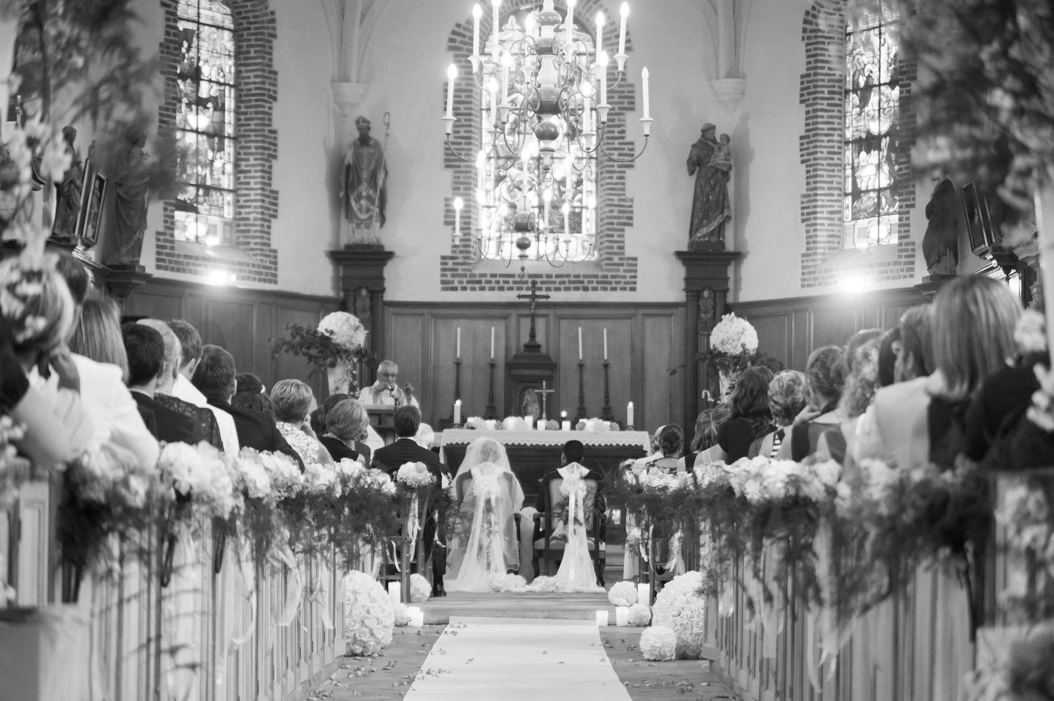 Black and white photo of an elegant church wedding ceremony with floral-lined aisle and candlelit altar.