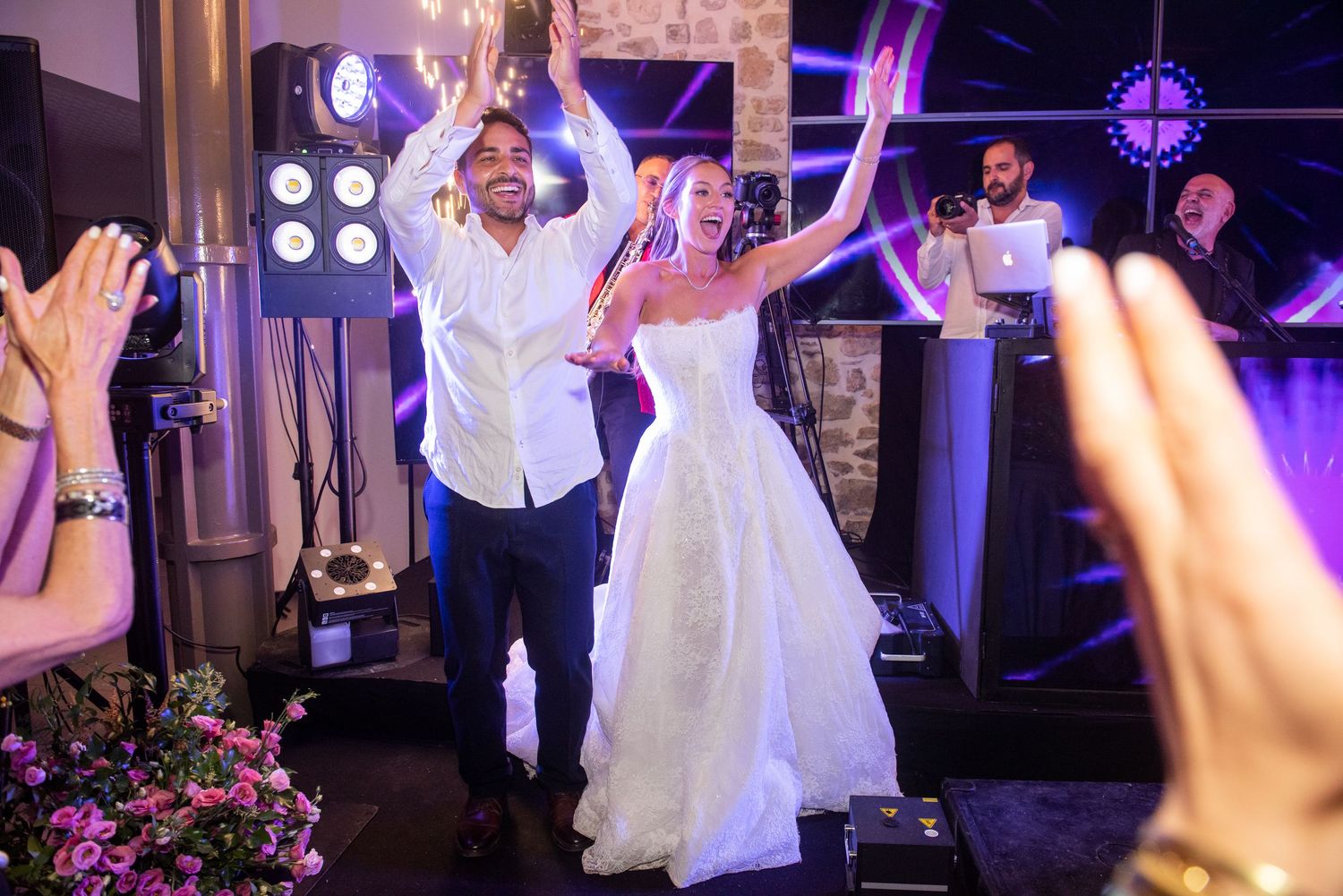A couple celebrates on a dance floor with raised arms during a wedding reception with purple lighting and floral decor.