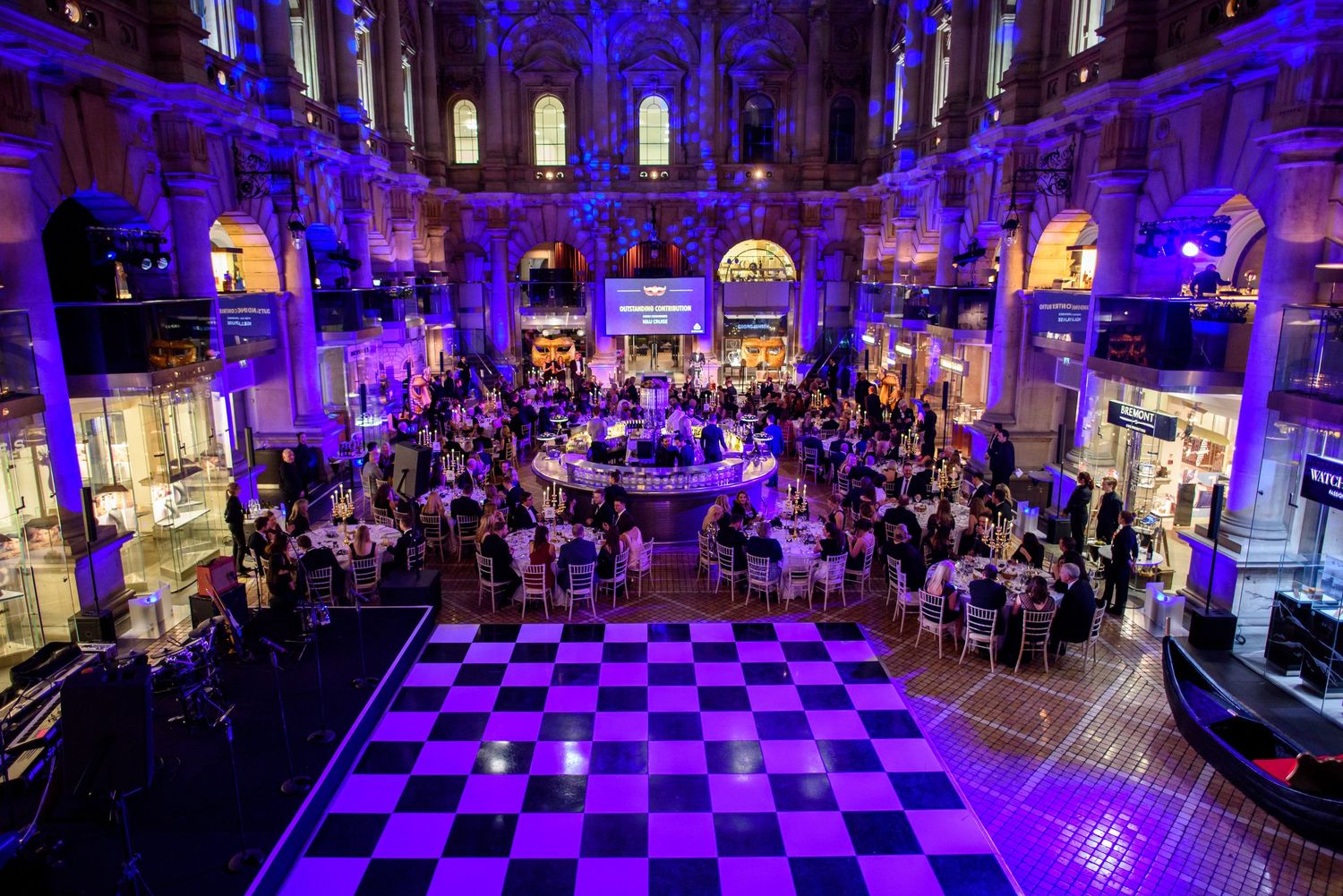 Grand ballroom with purple uplighting and checkered floor pattern filled with dining tables.