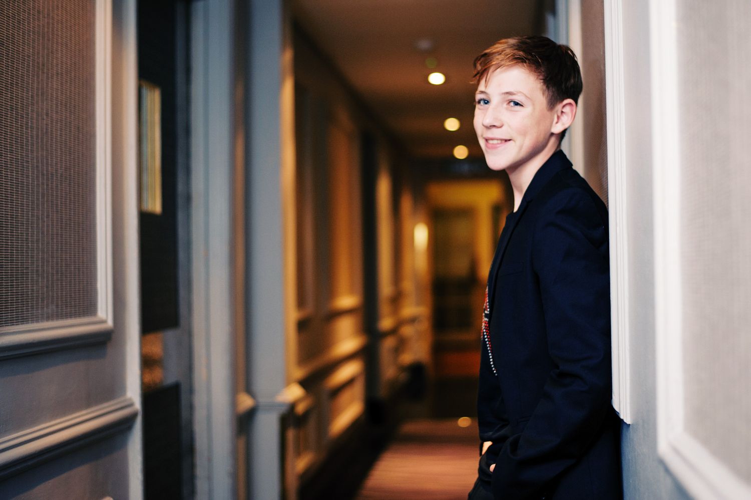 A person in a black blazer stands in a dimly lit hotel hallway with framed artwork on the walls.