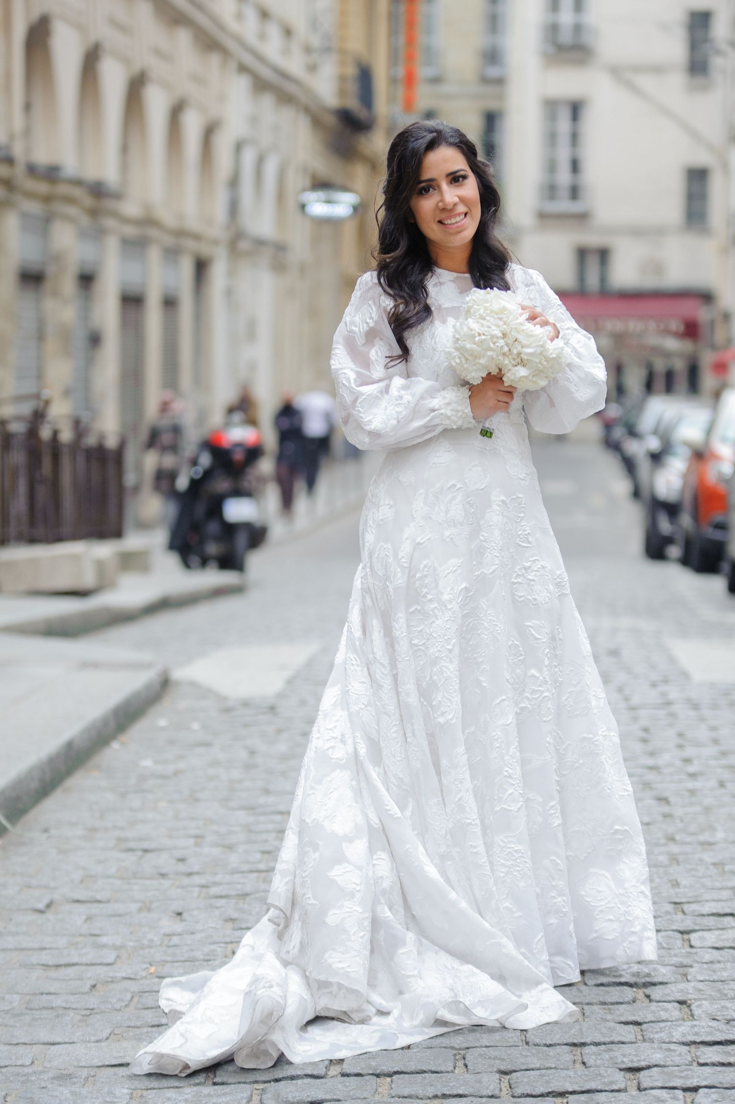 A person in a flowing white lace wedding dress poses gracefully on a cobblestone street in an elegant European setting.