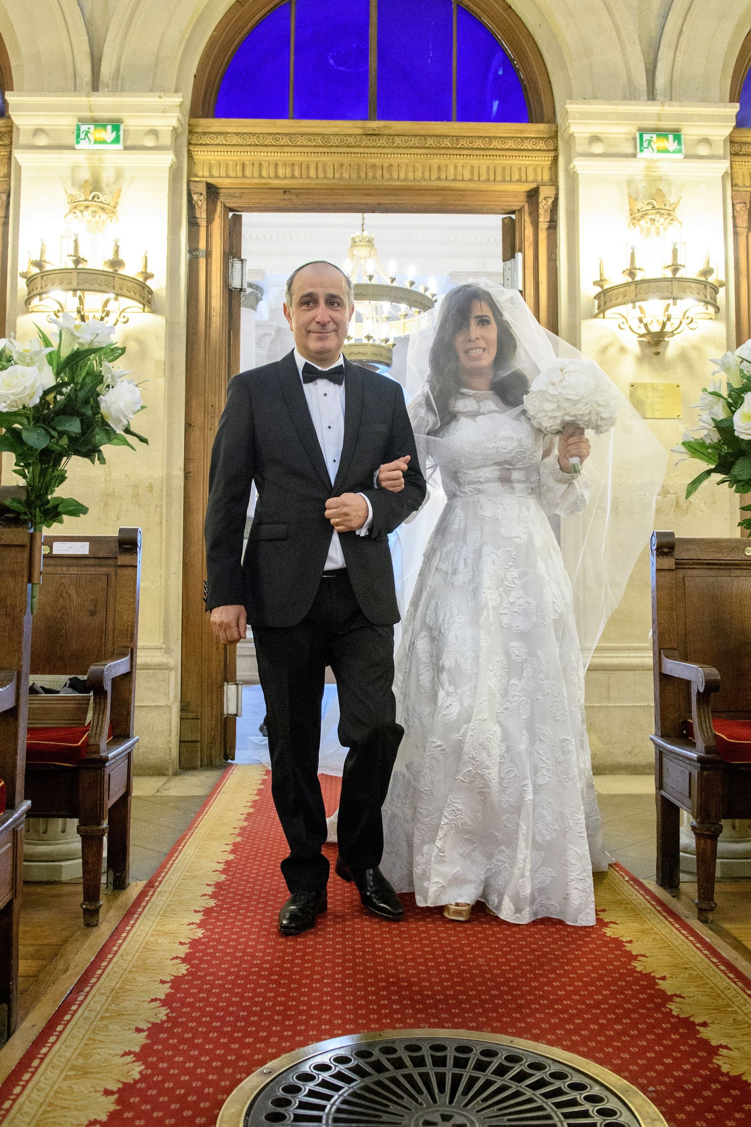 A couple walks down a red carpeted aisle in an elegant venue with ornate architectural details and lighting.
