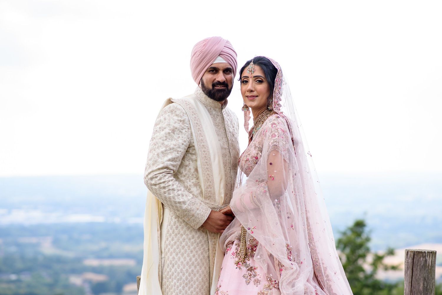 A couple in traditional Indian wedding attire poses outdoors with a scenic mountain view in the background.
