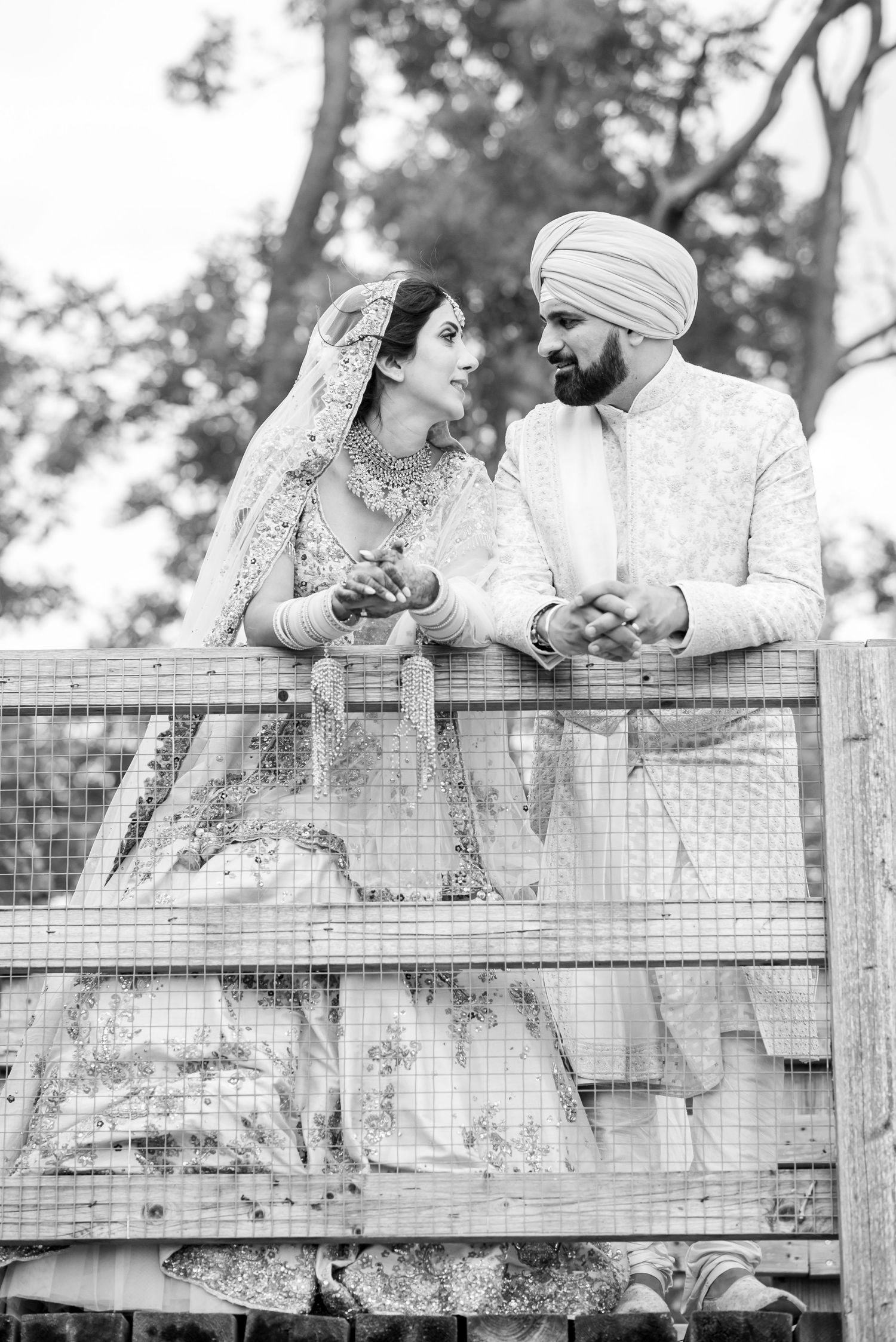 A black and white photo series shows a couple in traditional Indian wedding attire leaning on a wooden fence.