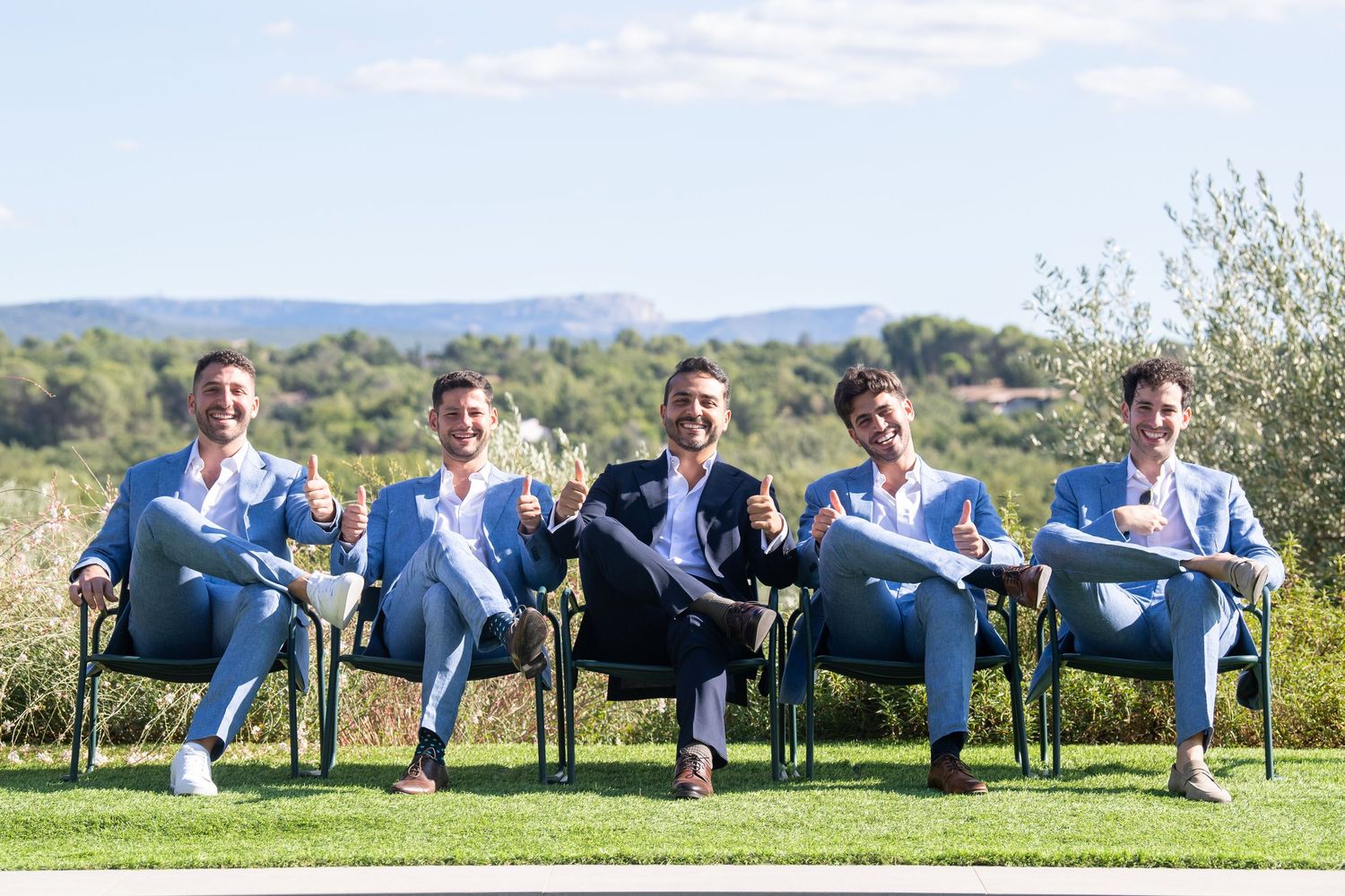 Five groomsmen in light blue suits sit outdoors in chairs against a scenic landscape backdrop.