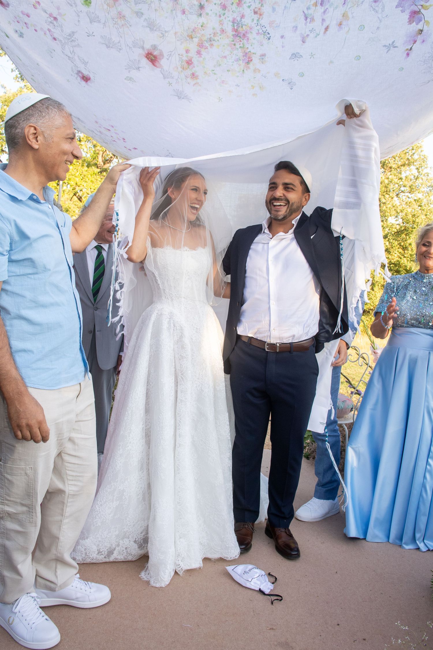 A joyful wedding celebration outdoors with guests holding a white cloth canopy over a bride in a flowing white gown.