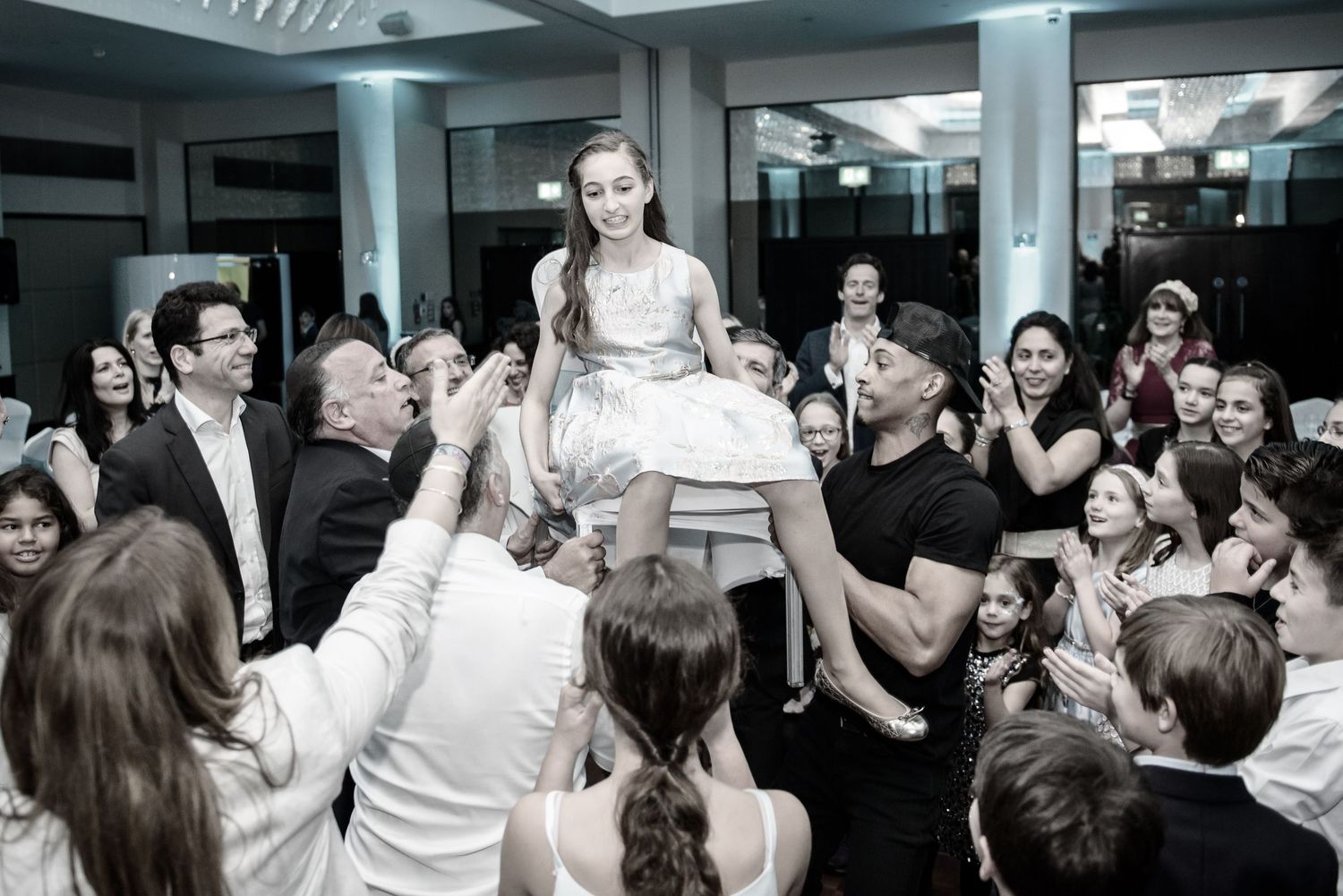 A crowd lifts someone wearing a white dress during a celebratory dance at an indoor reception event.
