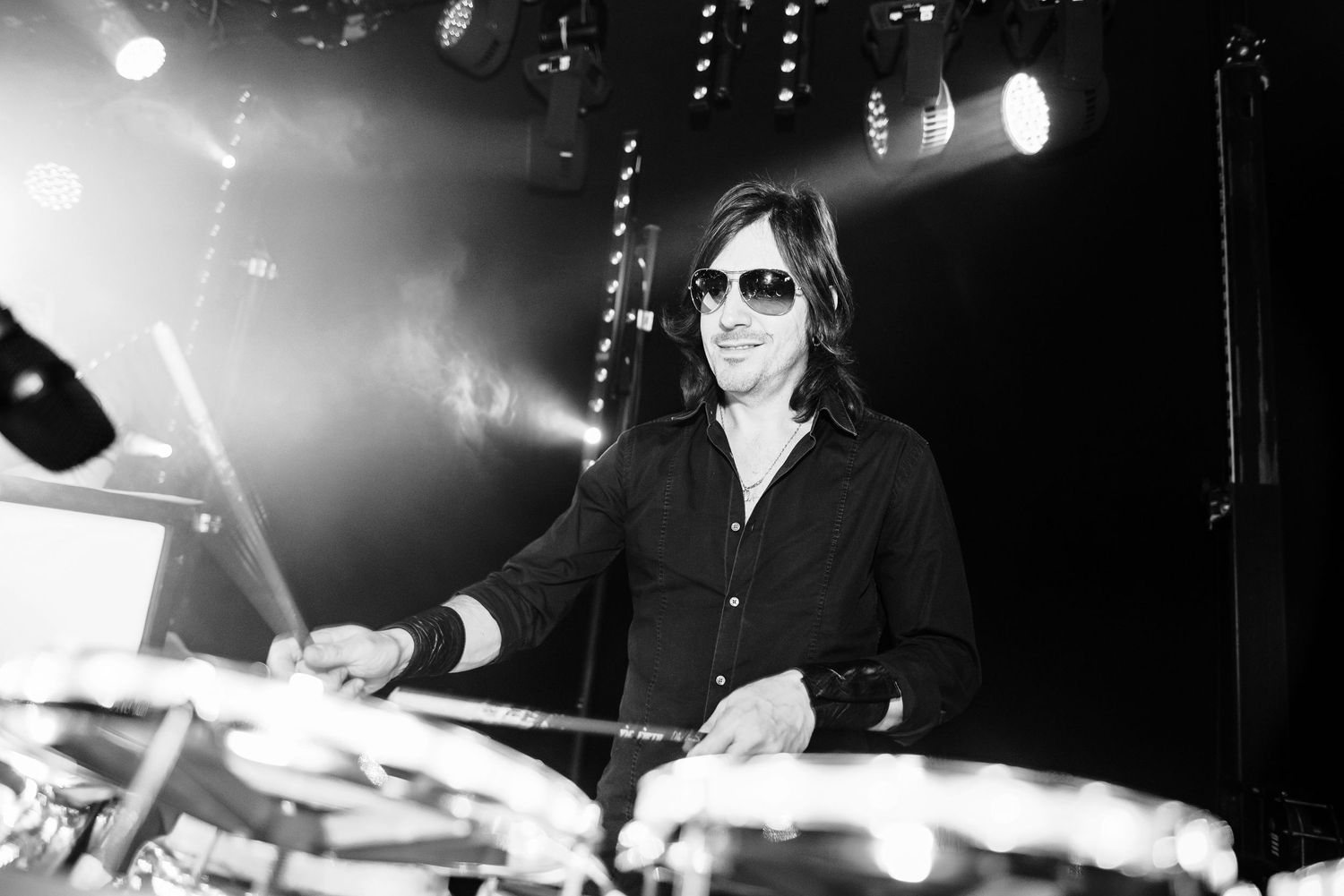 A drummer performs energetically on stage with dramatic lighting and smoke effects during a live concert performance.