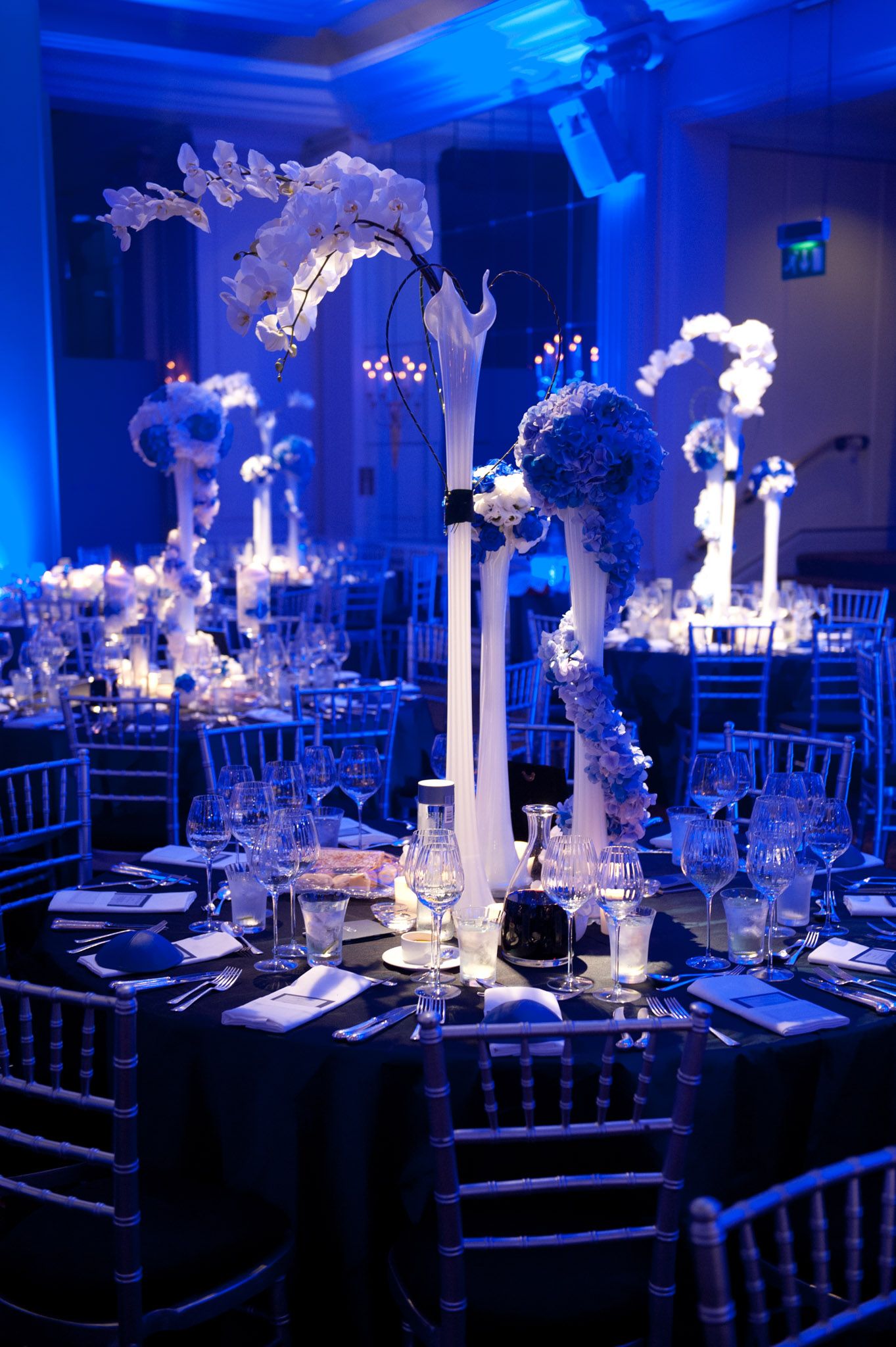 Elegant wedding reception tables illuminated in dramatic blue lighting with tall white floral centerpieces and crystal decor.