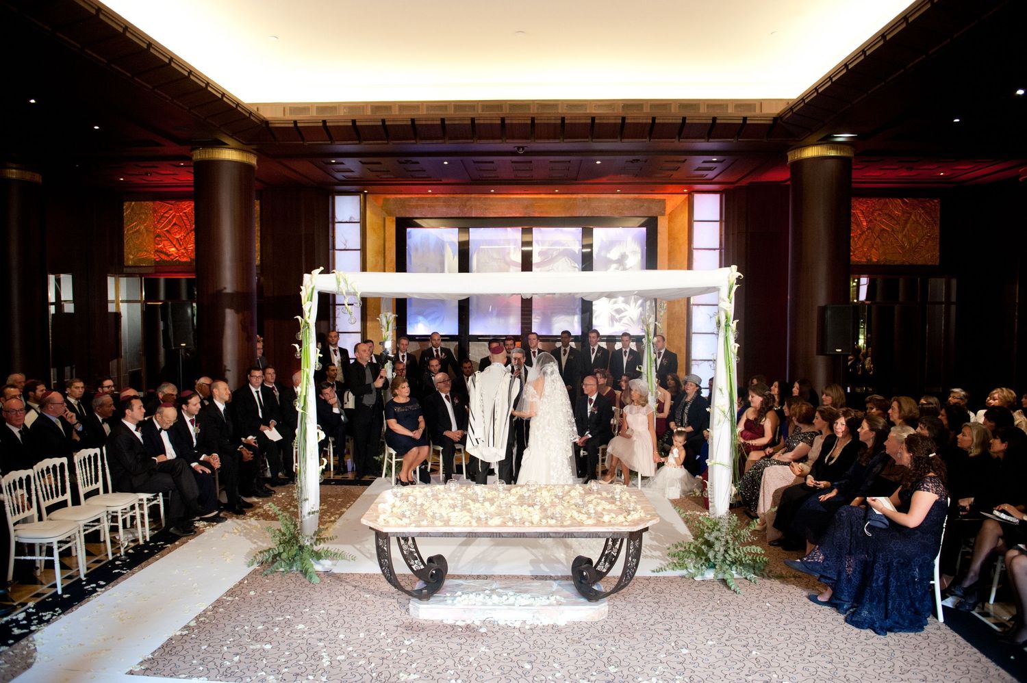 Wedding ceremony setup in an elegant ballroom with white chairs and decorative lighting along the aisle.