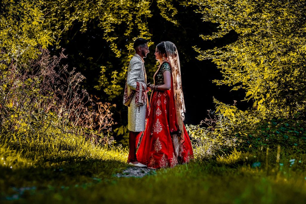 Couple in traditional Indian wedding attire standing together in a lush green garden with sunlit trees in the background.