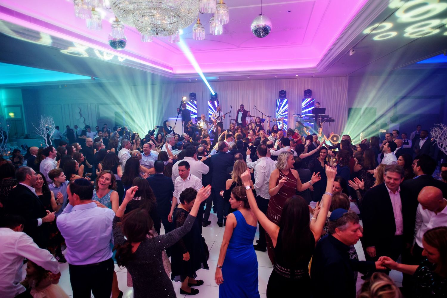 A crowded dance floor at a wedding reception illuminated by colorful pink and blue lighting effects.