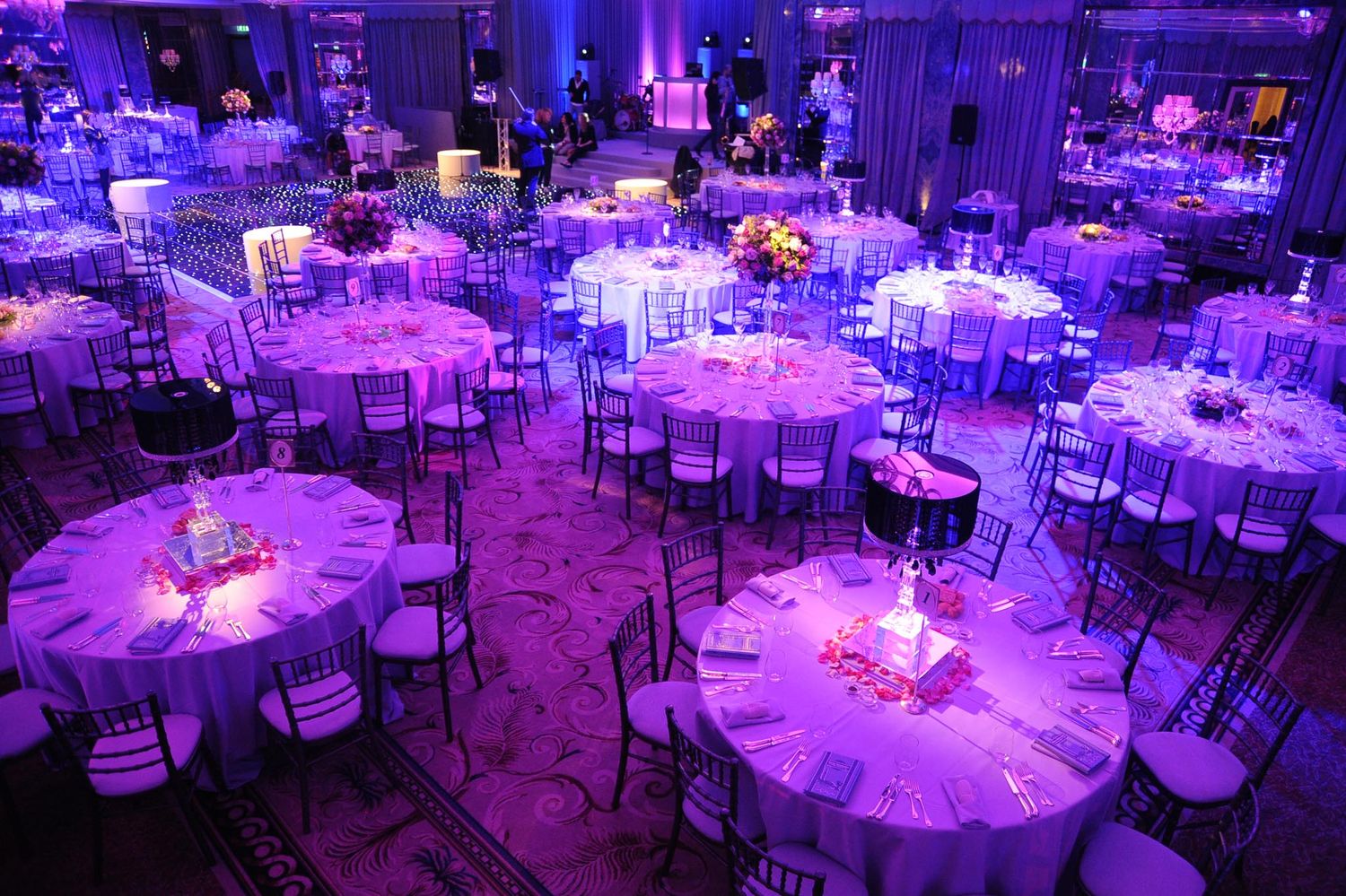 Round banquet tables illuminated by purple uplighting create an elegant atmosphere at an indoor wedding reception venue.