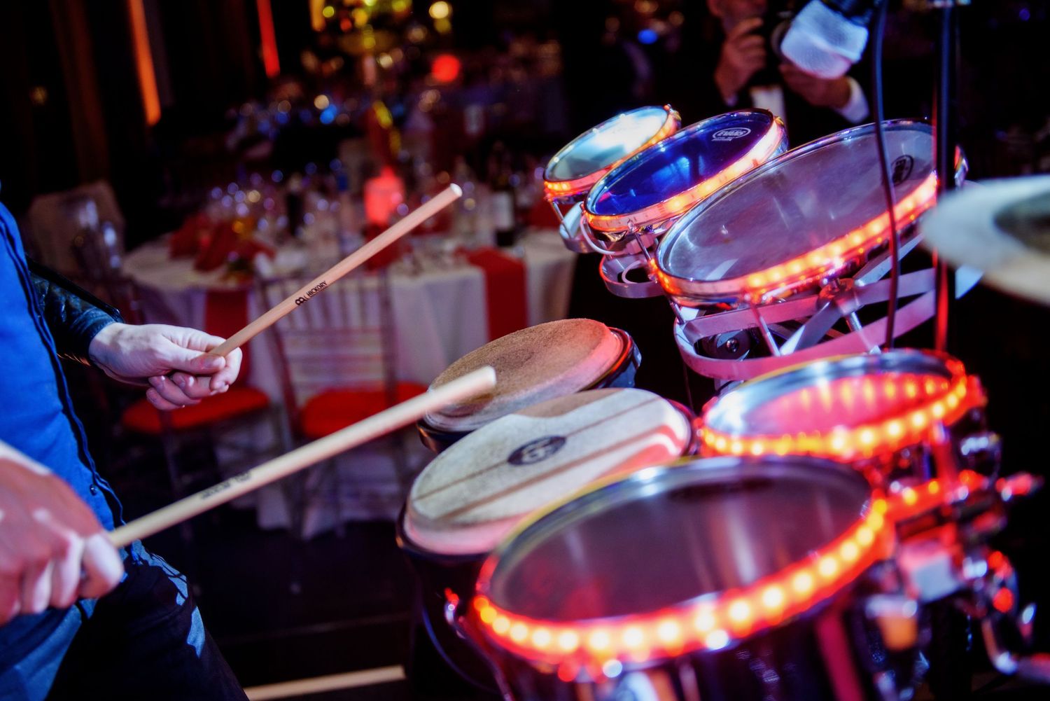 Illuminated drum set with red LED lights glowing in dark concert setting while drummer performs with drumsticks.