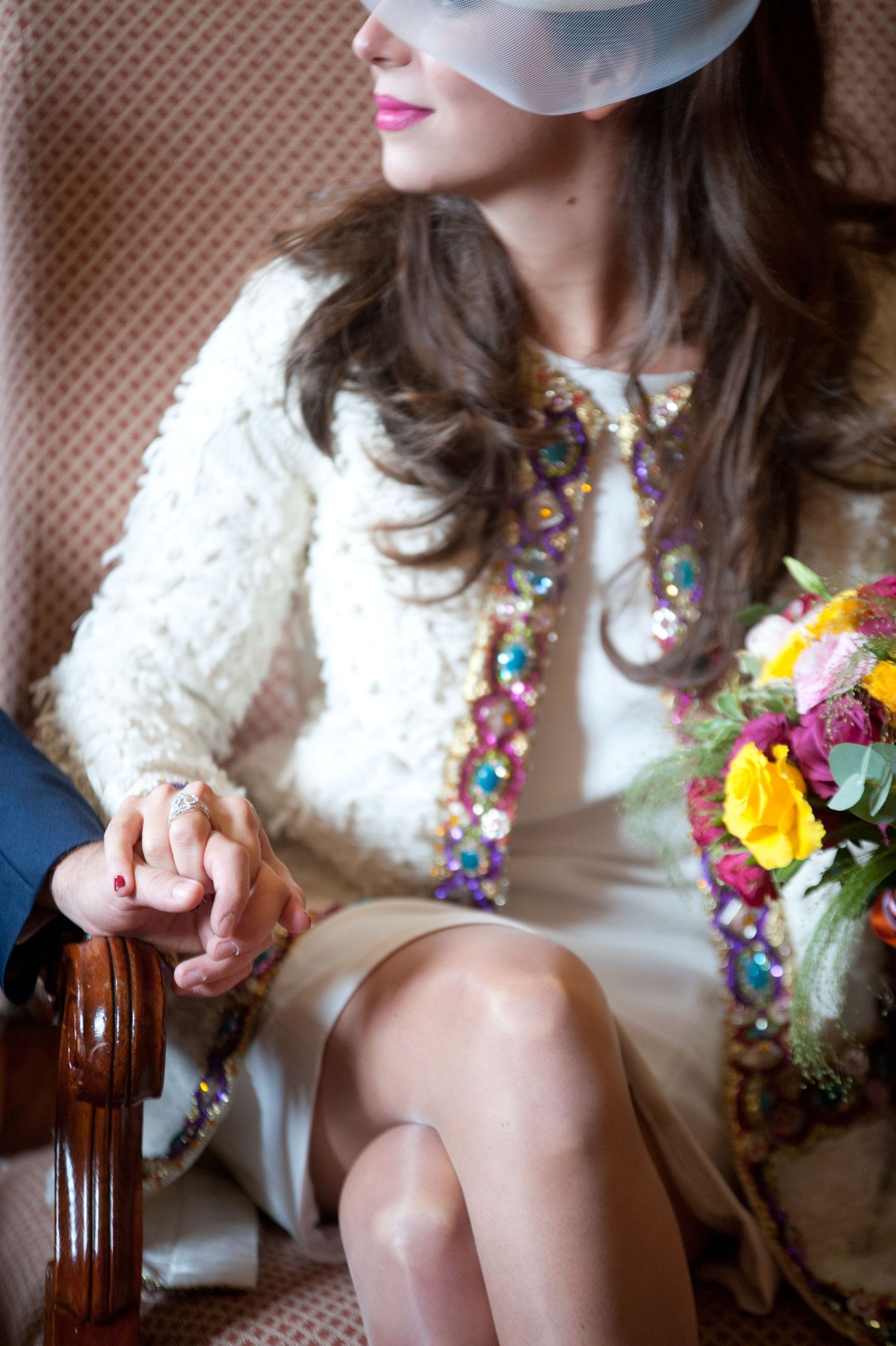 A person in a white dress sits on a wooden chair wearing colorful beaded jewelry and holding bright flowers.