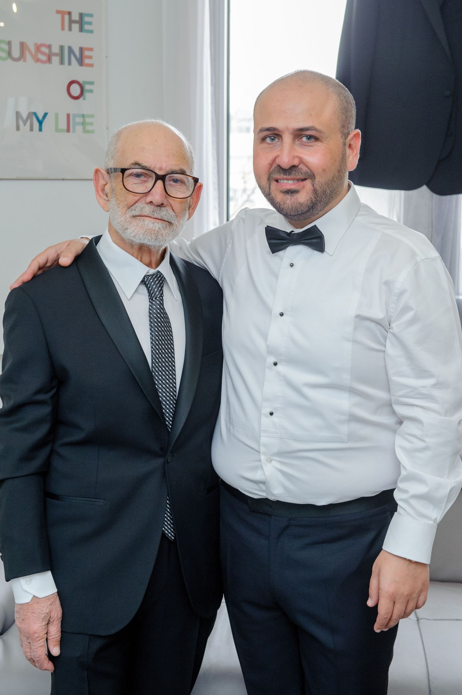 Two men in formal attire, one wearing a black suit and one in white dress shirt, pose together for photos.