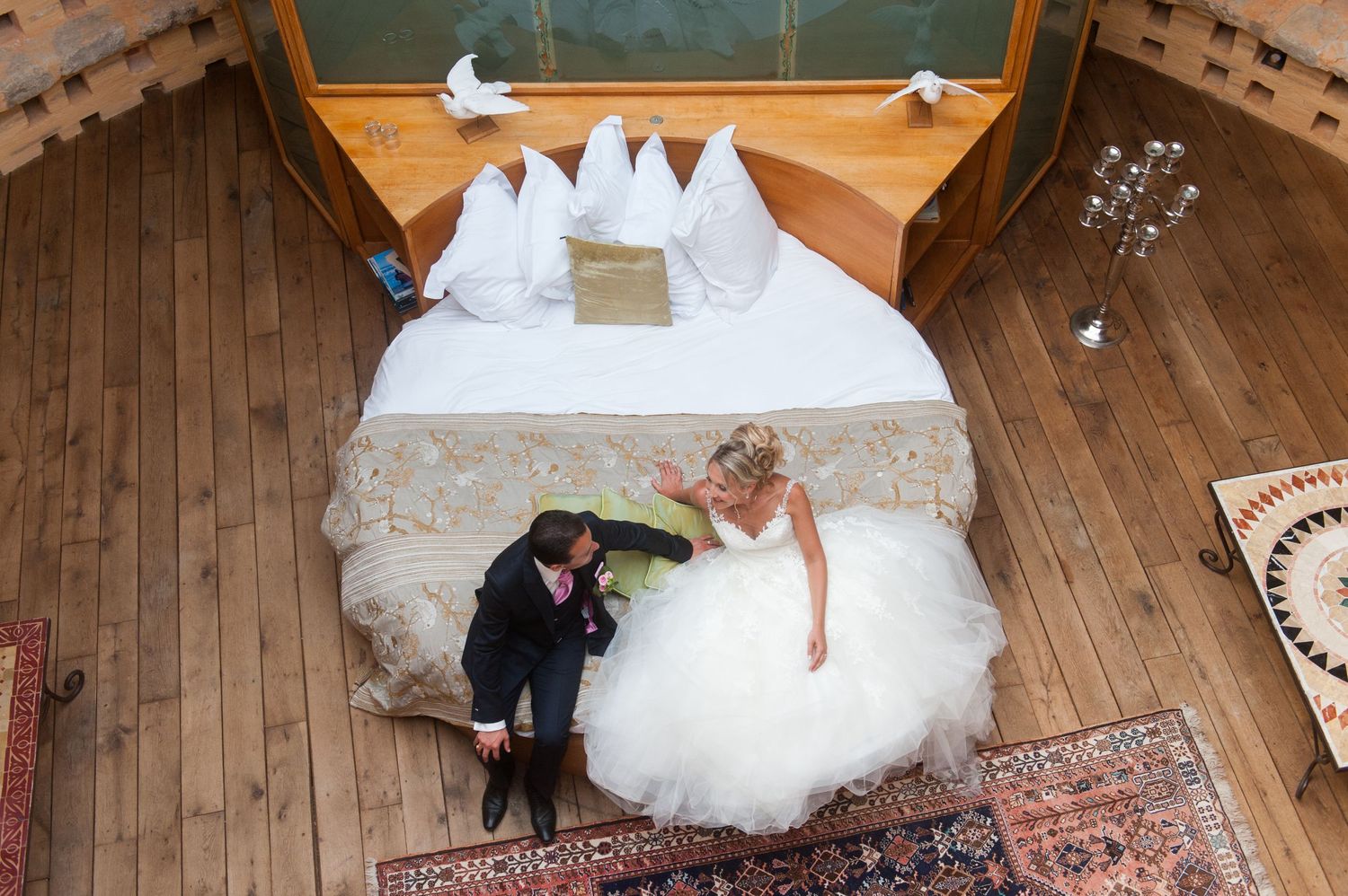 Aerial view of bride and groom sharing a romantic moment beside a large white bed with dove decorations.