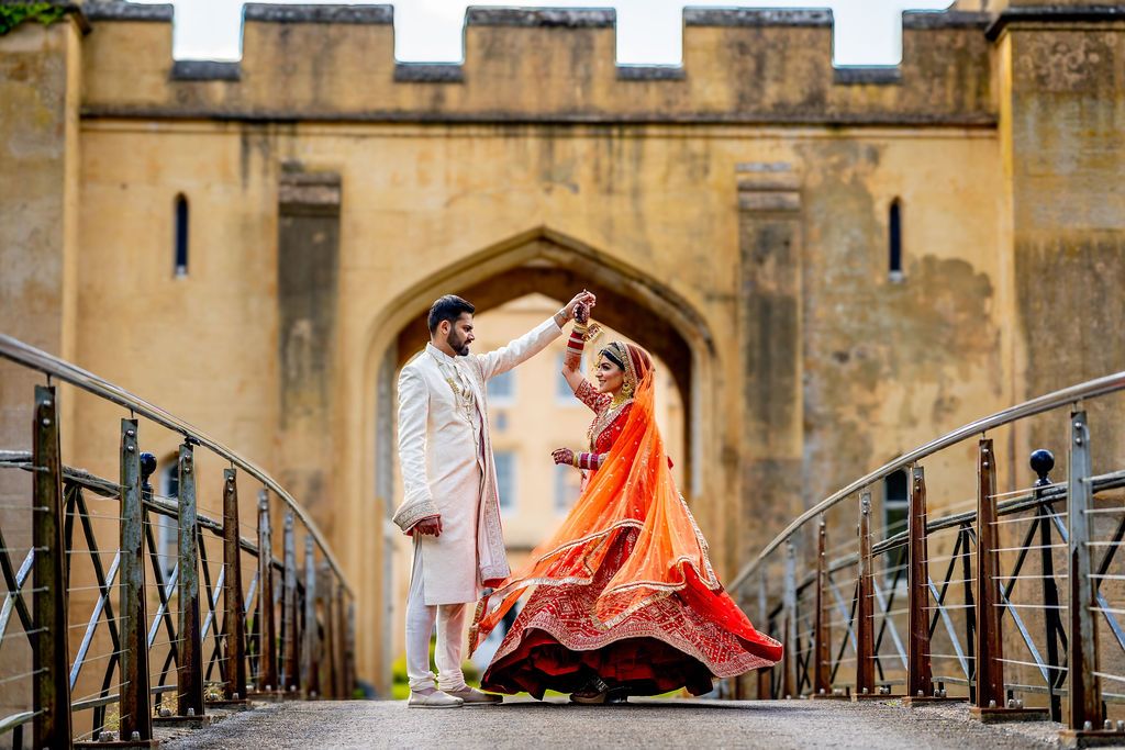 Indian couple in traditional wedding attire dancing on a bridge, bride in orange lehenga, groom in white sherwani.