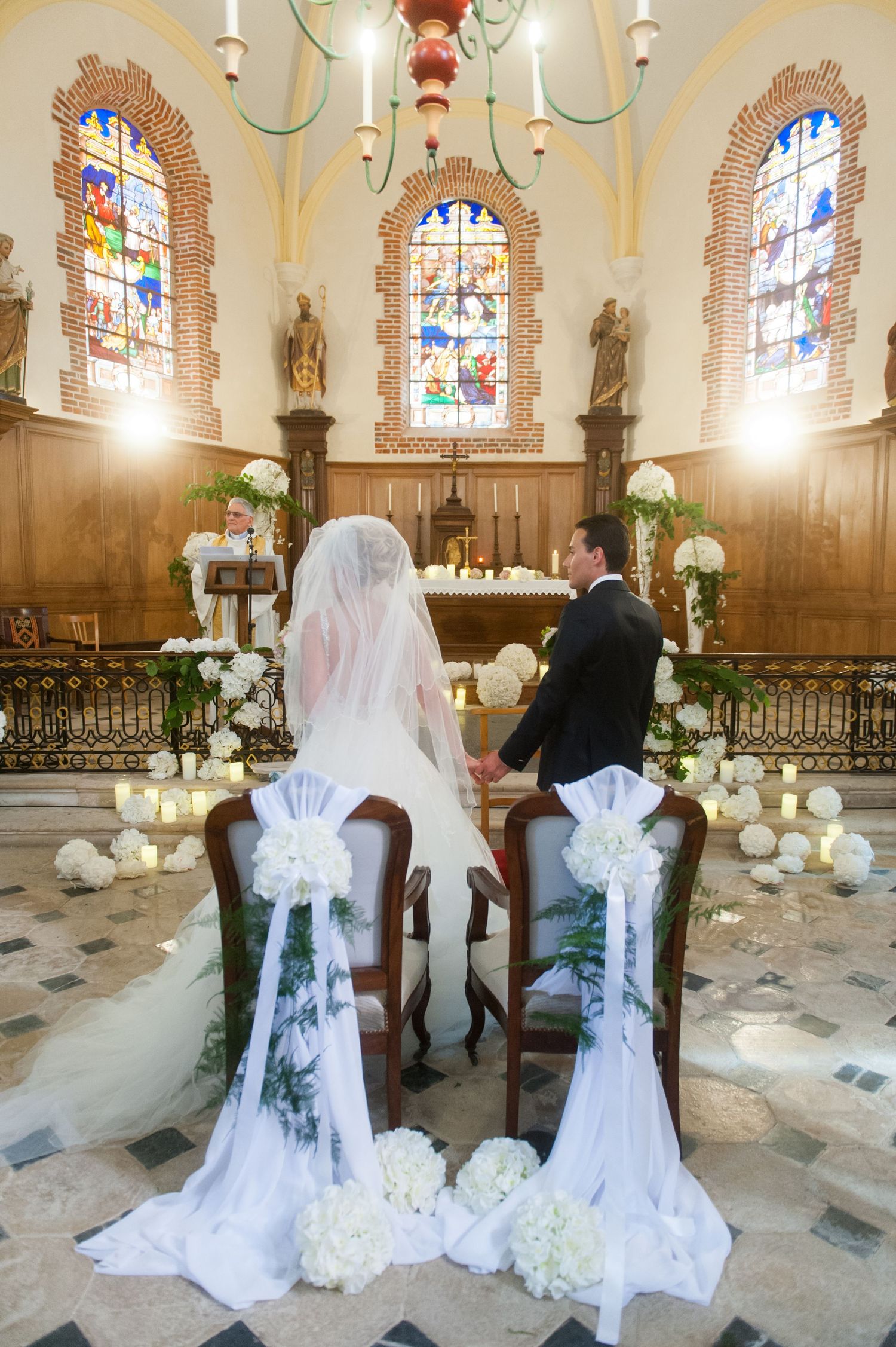 Bride and groom seated at church altar during wedding ceremony with white floral decorations and stained glass windows.