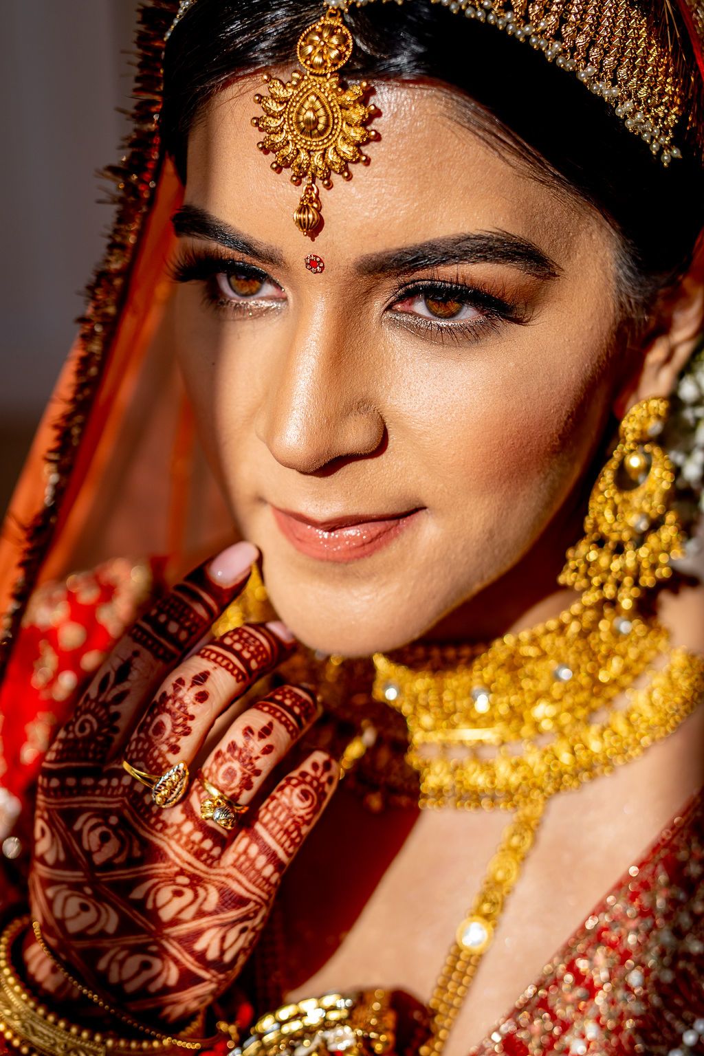 Indian bride in red dupatta with gold jewelry, maang tikka, and intricate mehndi henna on hands.