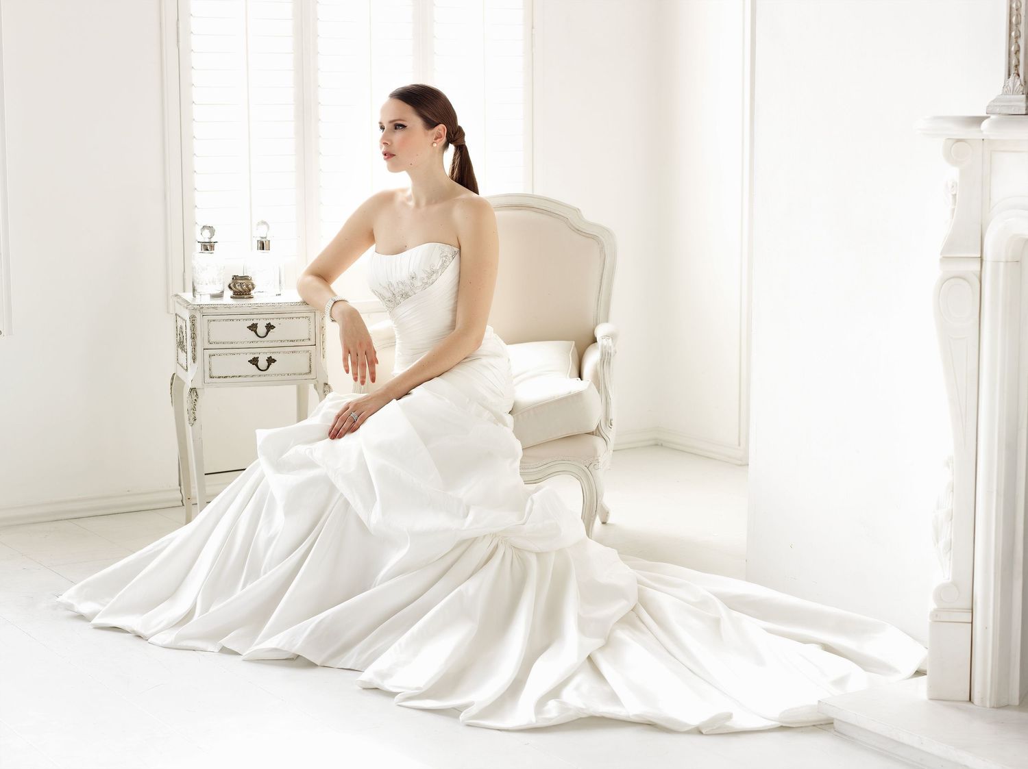 Bride in elegant strapless white wedding gown seated gracefully in a vintage chair in a bright, airy white room.