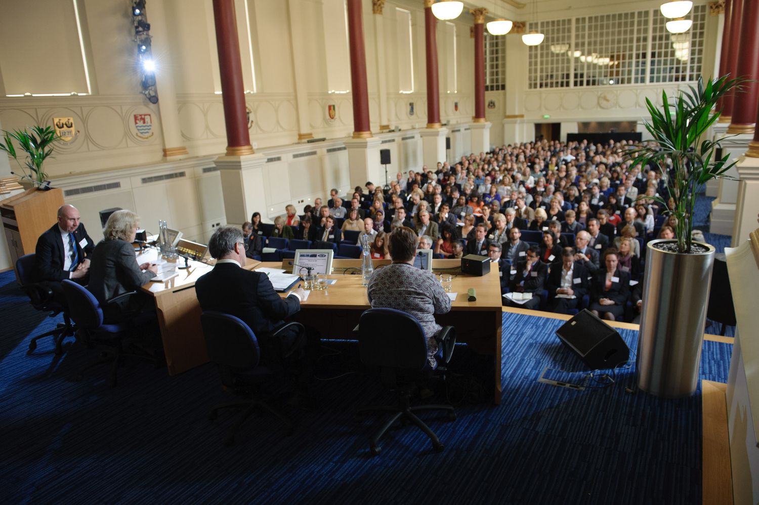 Large audience gathered in an auditorium for a conference or presentation with speakers at the front.