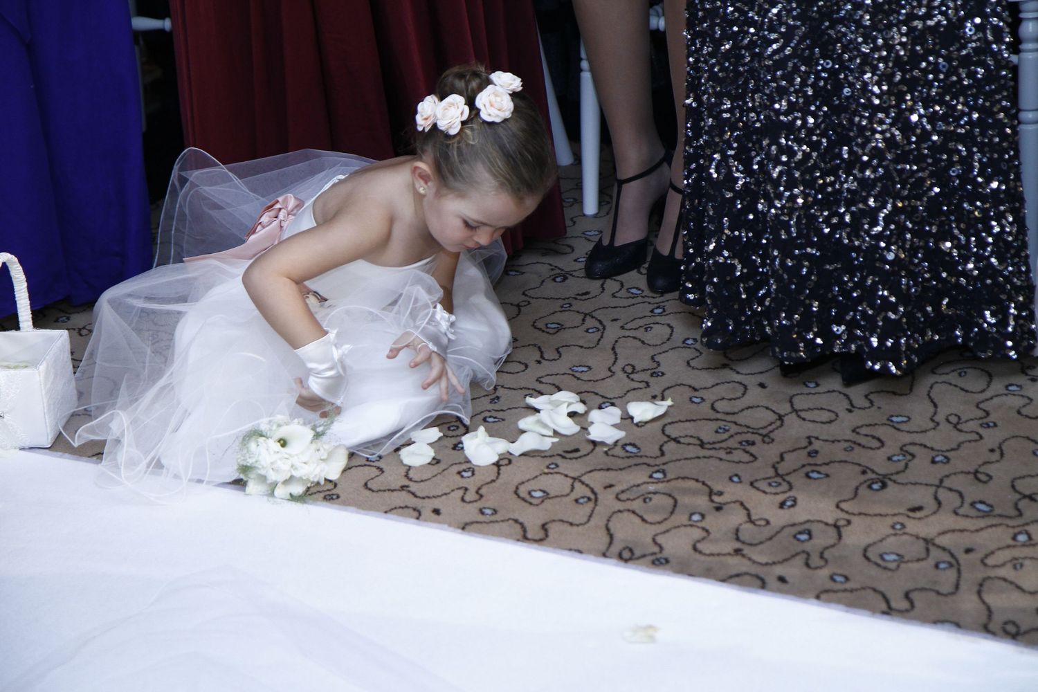 A bride in a white wedding dress lies gracefully on a patterned carpet surrounded by scattered rose petals.