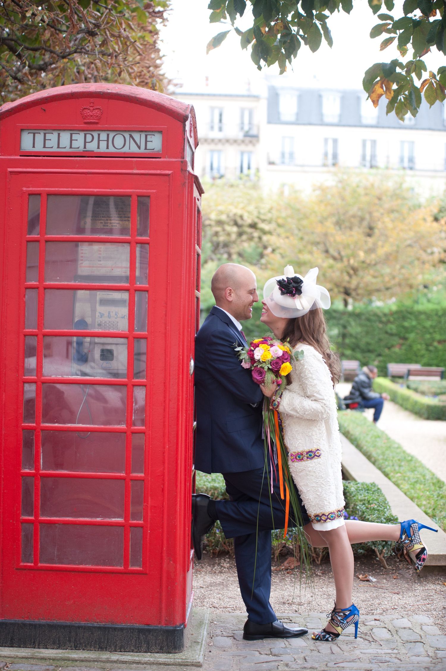A romantic couple poses near iconic red British telephone boxes in a scenic park setting.