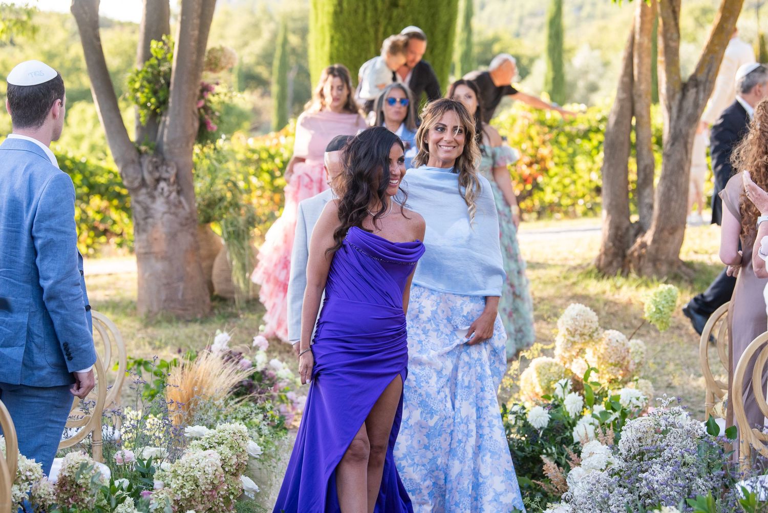 A group gathers in a sunlit garden setting with white flowers and trees creating a beautiful outdoor atmosphere.