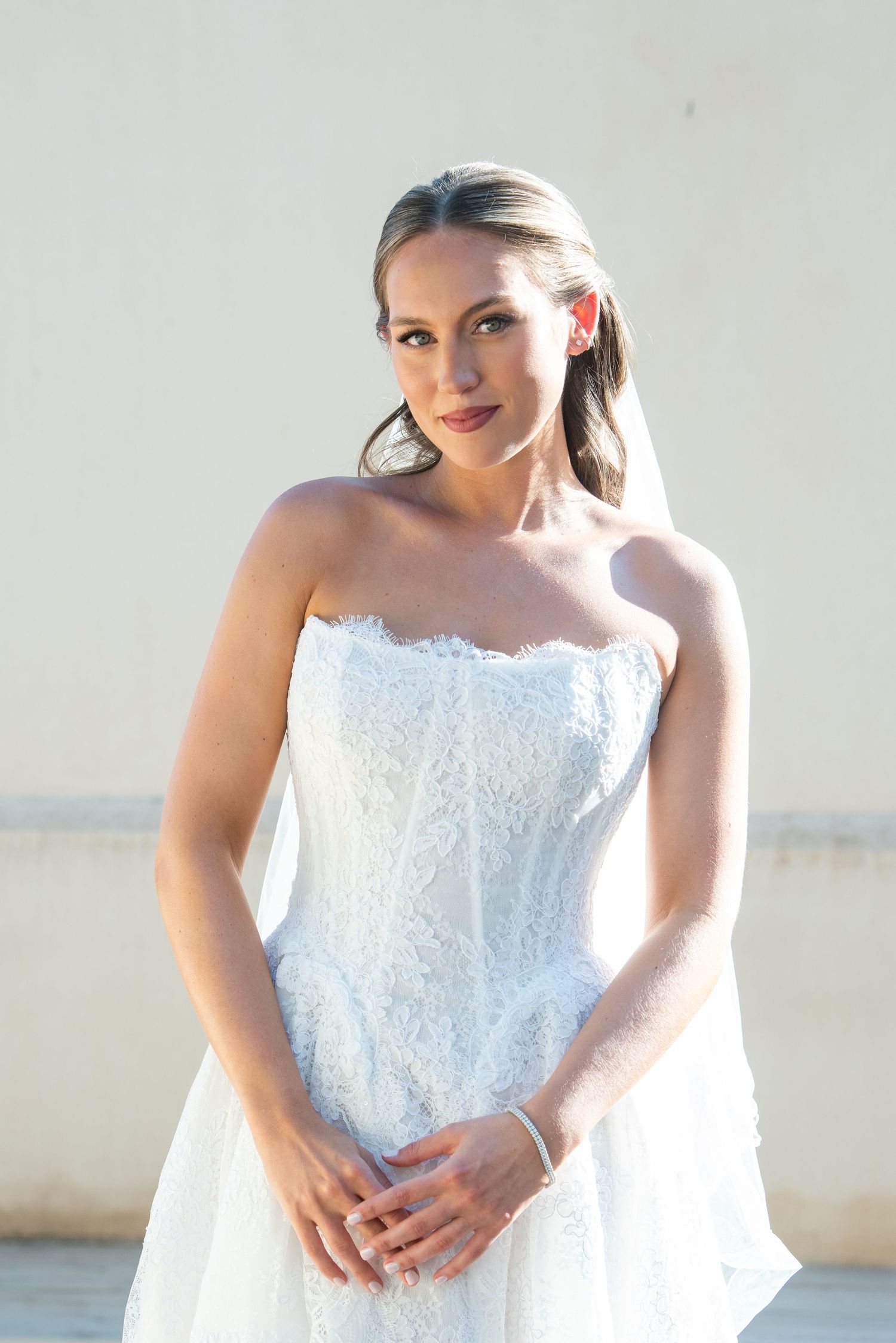 Bride poses in a strapless white lace wedding gown against a light colored wall.