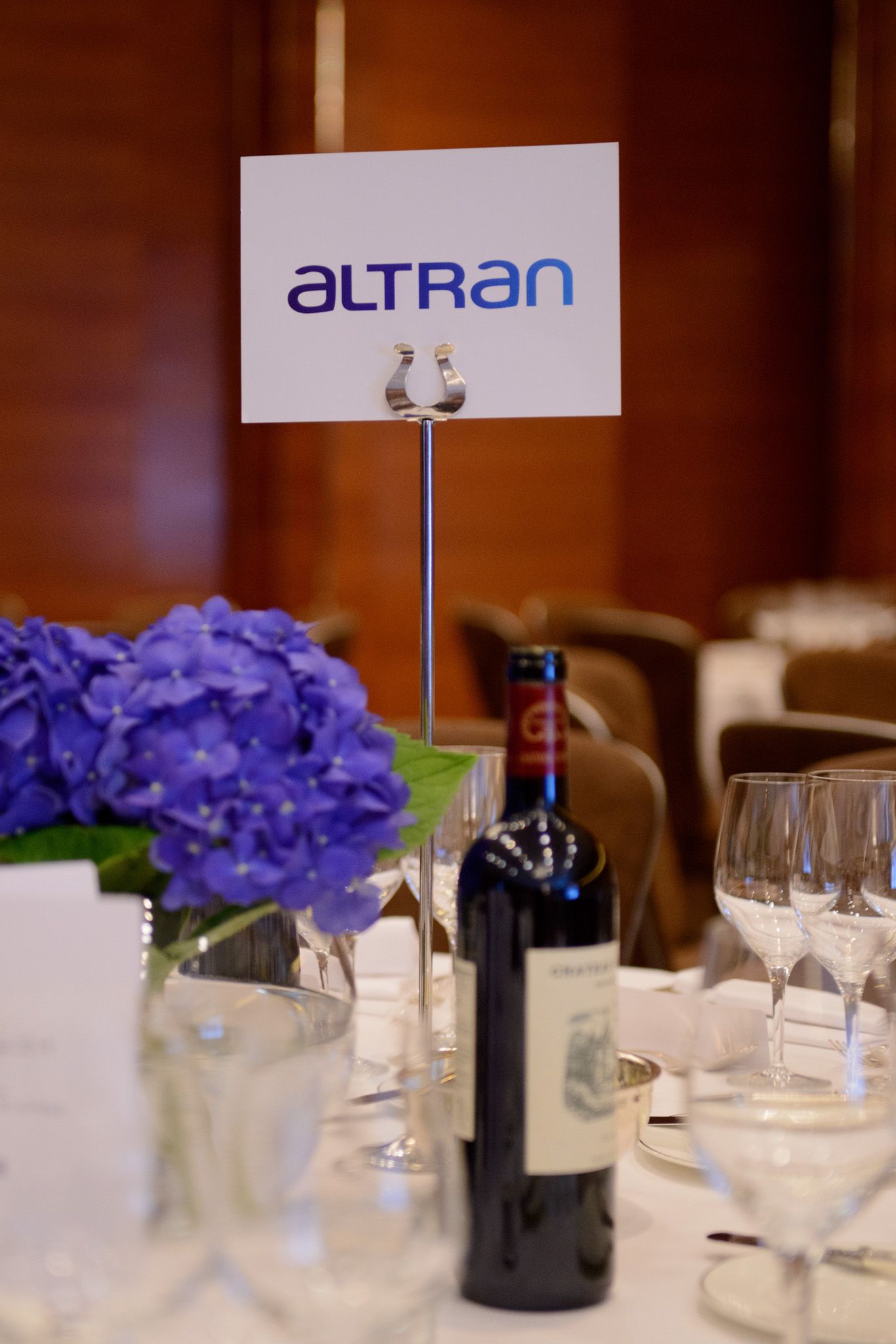 Elegant table setting with Altran company sign, wine bottles, and purple hydrangeas as centerpiece.