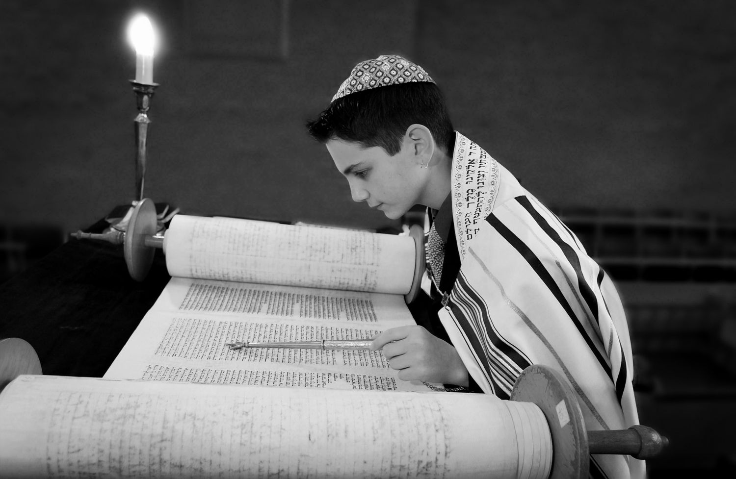 Person wearing a prayer shawl reads from a Torah scroll in black and white.