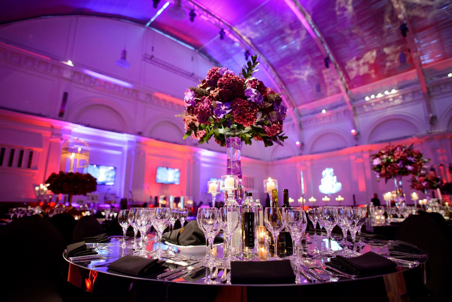Wedding reception venue decorated with crystal chandeliers and bathed in vibrant purple and pink lighting.
