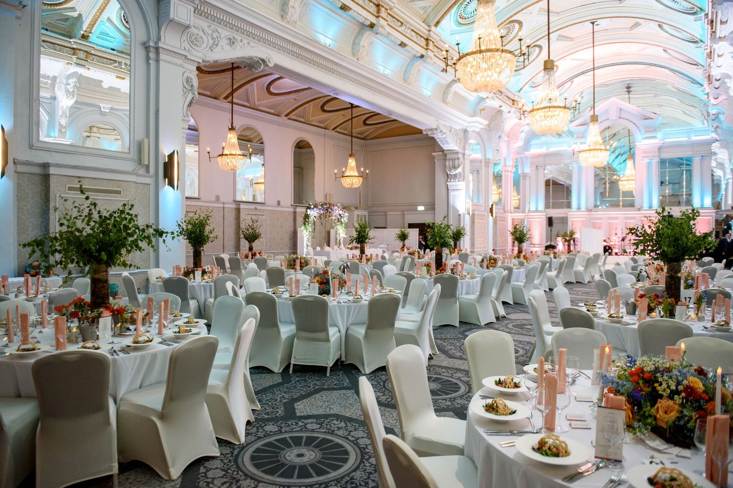 Elegant wedding reception venue with white chairs, hanging lights, and floral centerpieces in a bright indoor space.