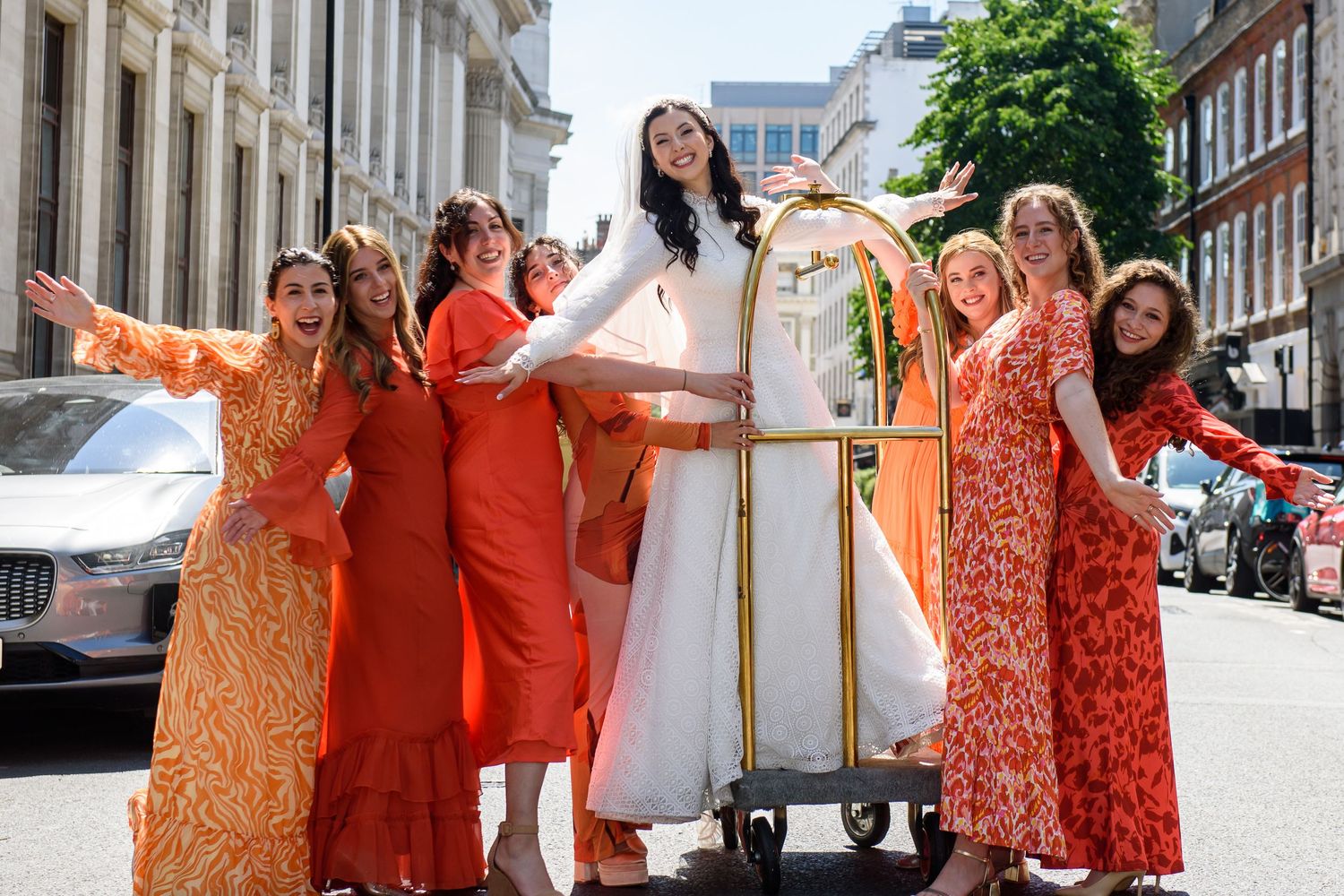 A group of bridesmaids in vibrant orange and floral dresses pose together on a city street during a wedding celebration.