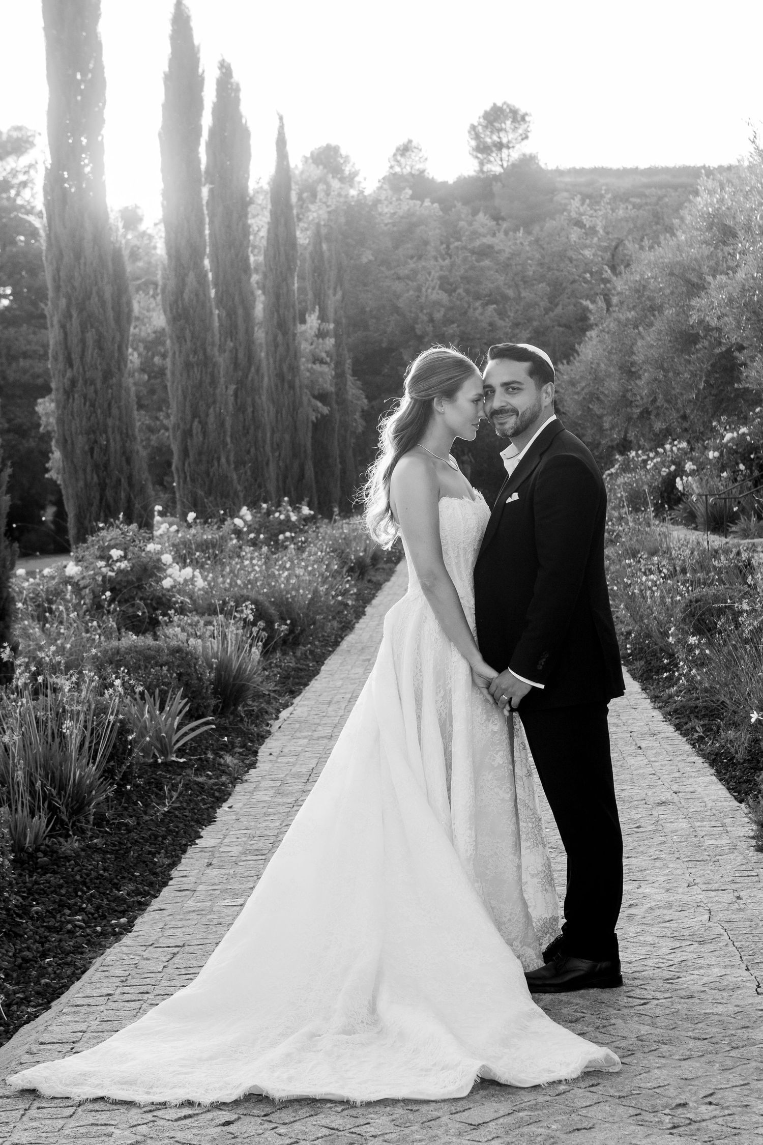A romantic black and white wedding photo on a garden path lined with tall cypress trees and blooming flowers.